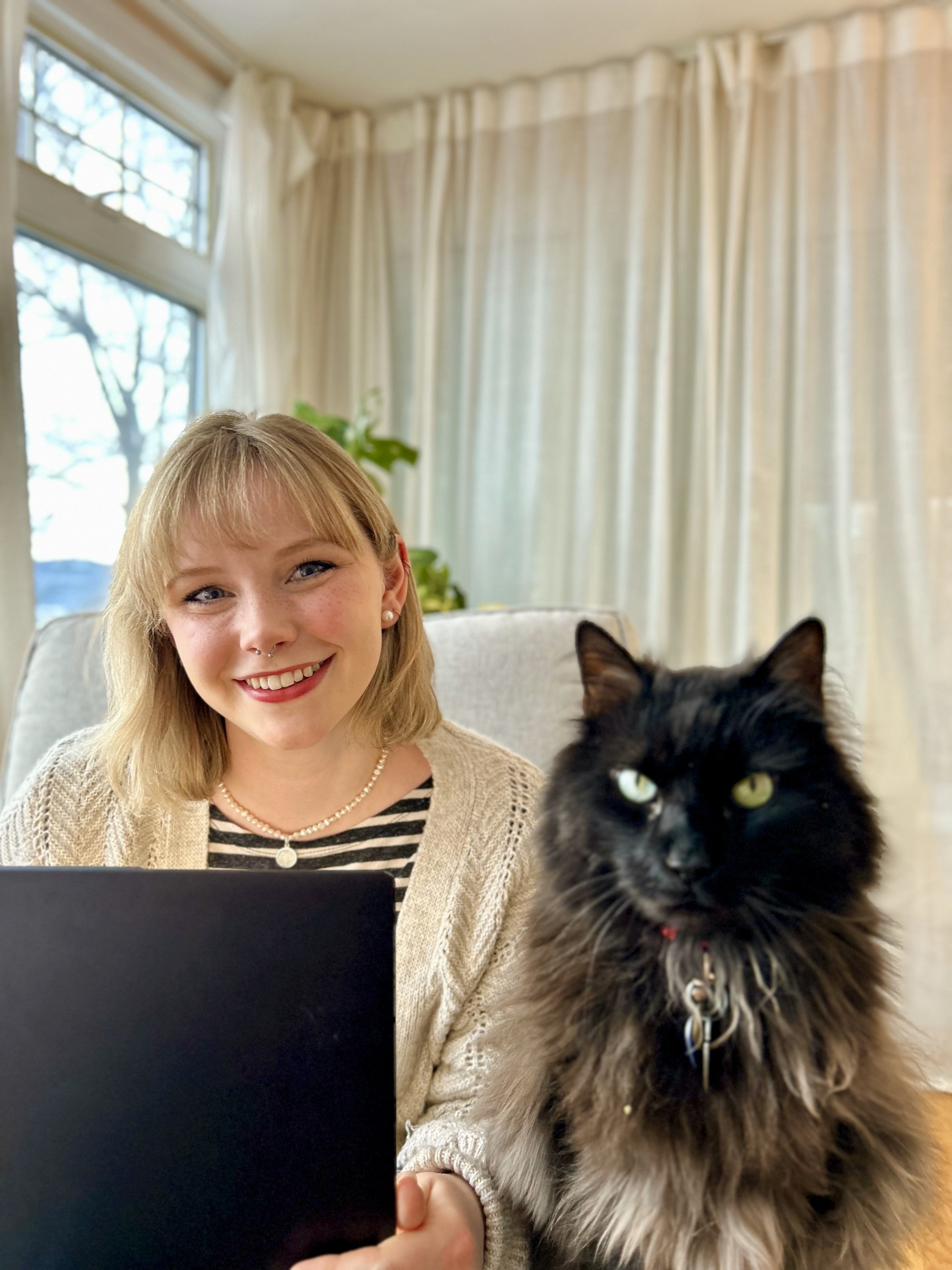 A woman with blonde hair and a black and white striped shirt smiling at the camera, sitting at a laptop with a black and brown long-haired cat sitting next to her. The room has cream-colored curtains and a window in the background.