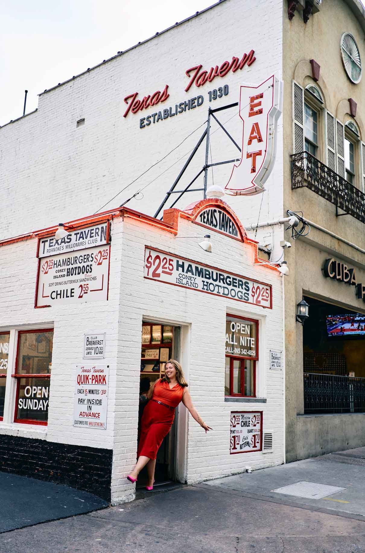 A woman in a red dress and pink shoes standing at the entrance of Texas Tavern, a white brick building with vintage signs advertising burgers and hot dogs, and a large Texas Tavern neon sign on the roof.
