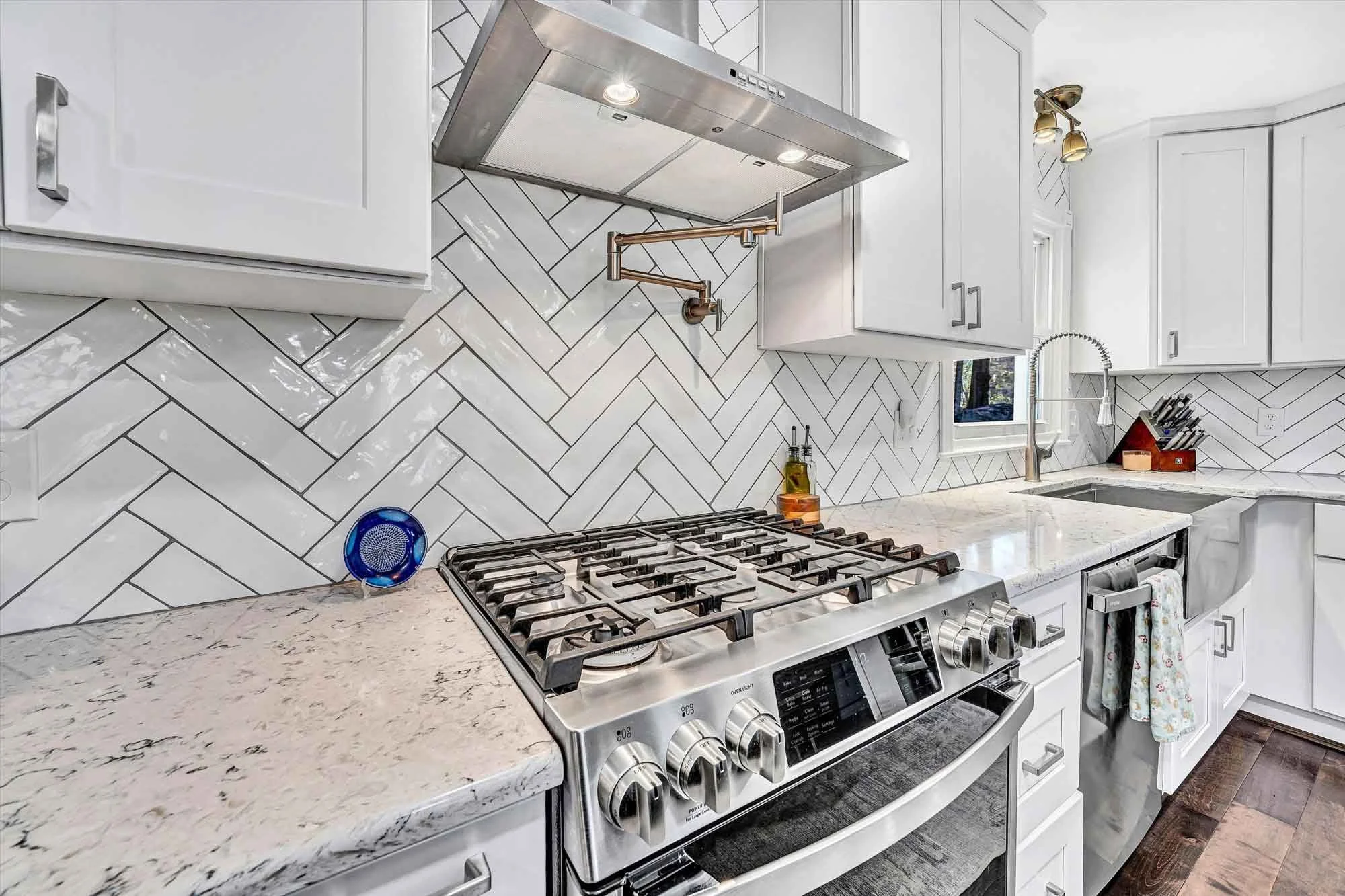 Modern kitchen with white cabinetry, a marble countertop, a stainless steel gas stove, a white tile backsplash in a herringbone pattern, a small window above the sink, and various kitchen accessories.