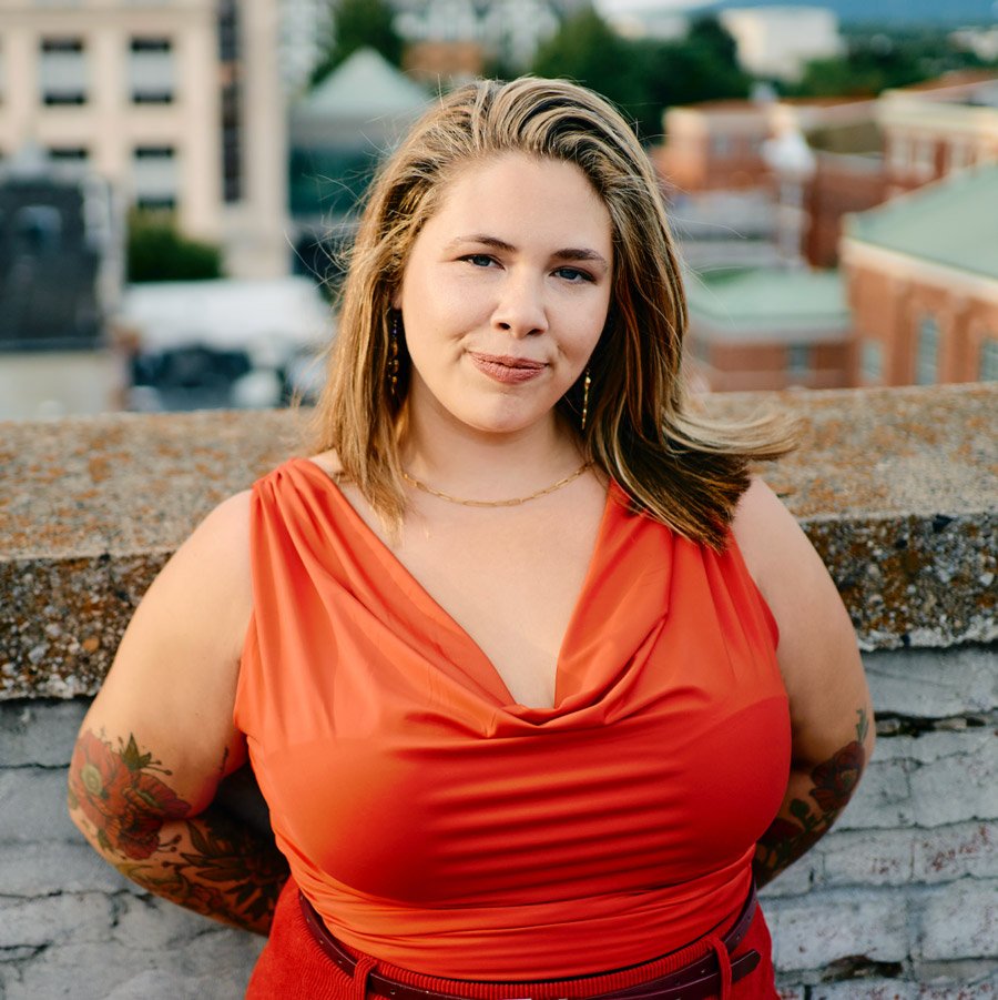 A young woman in an orange dress standing outdoors on a rooftop with city buildings in the background.