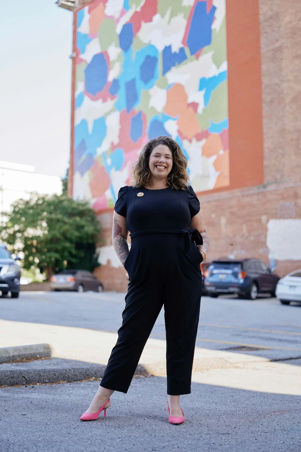 A woman in a black jumpsuit and pink high heels standing outdoors in front of a colorful mural wall, smiling with her hands in her pockets.