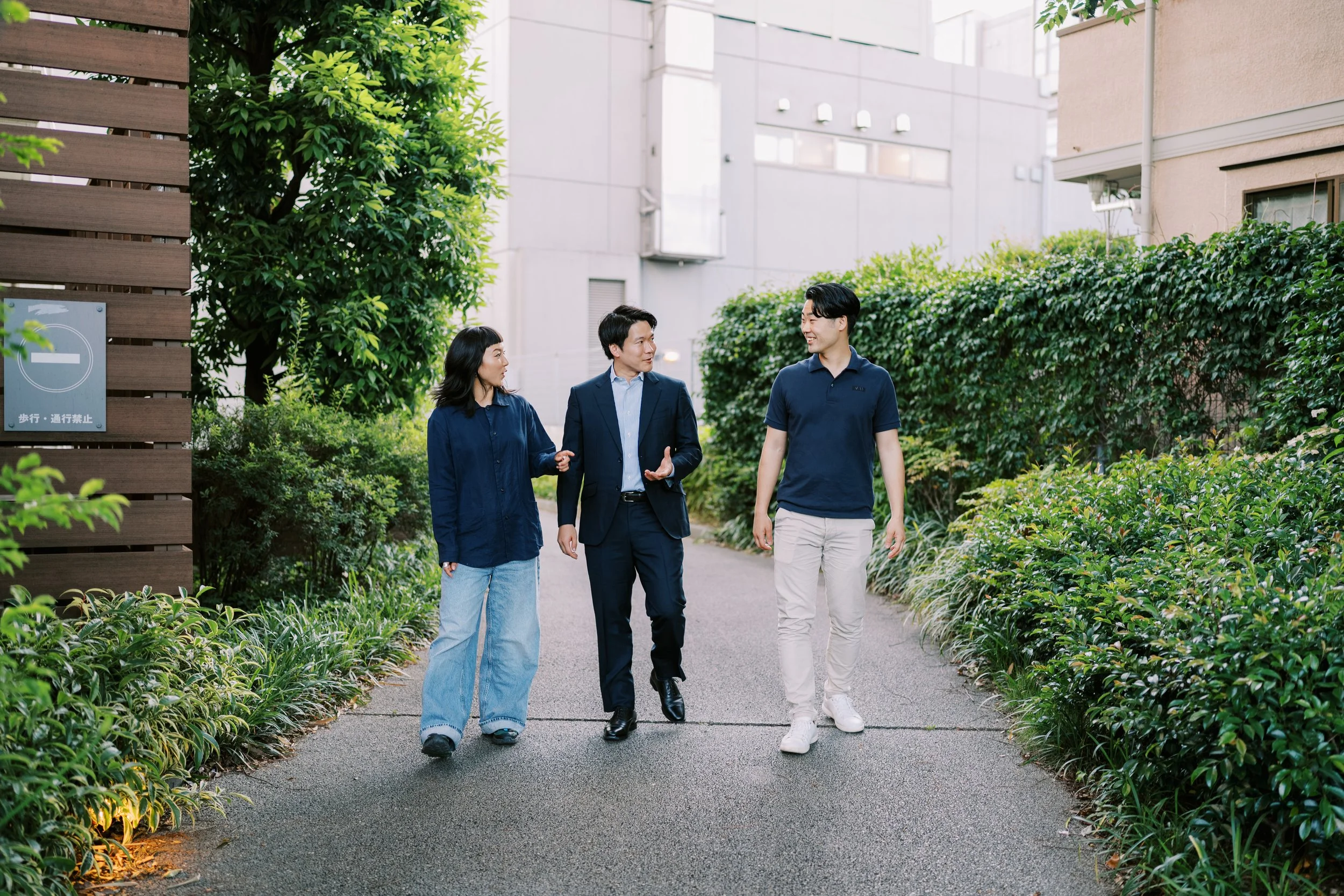 3人が緑豊かな公園を歩きながら会話している。背景には木と建物が見える。