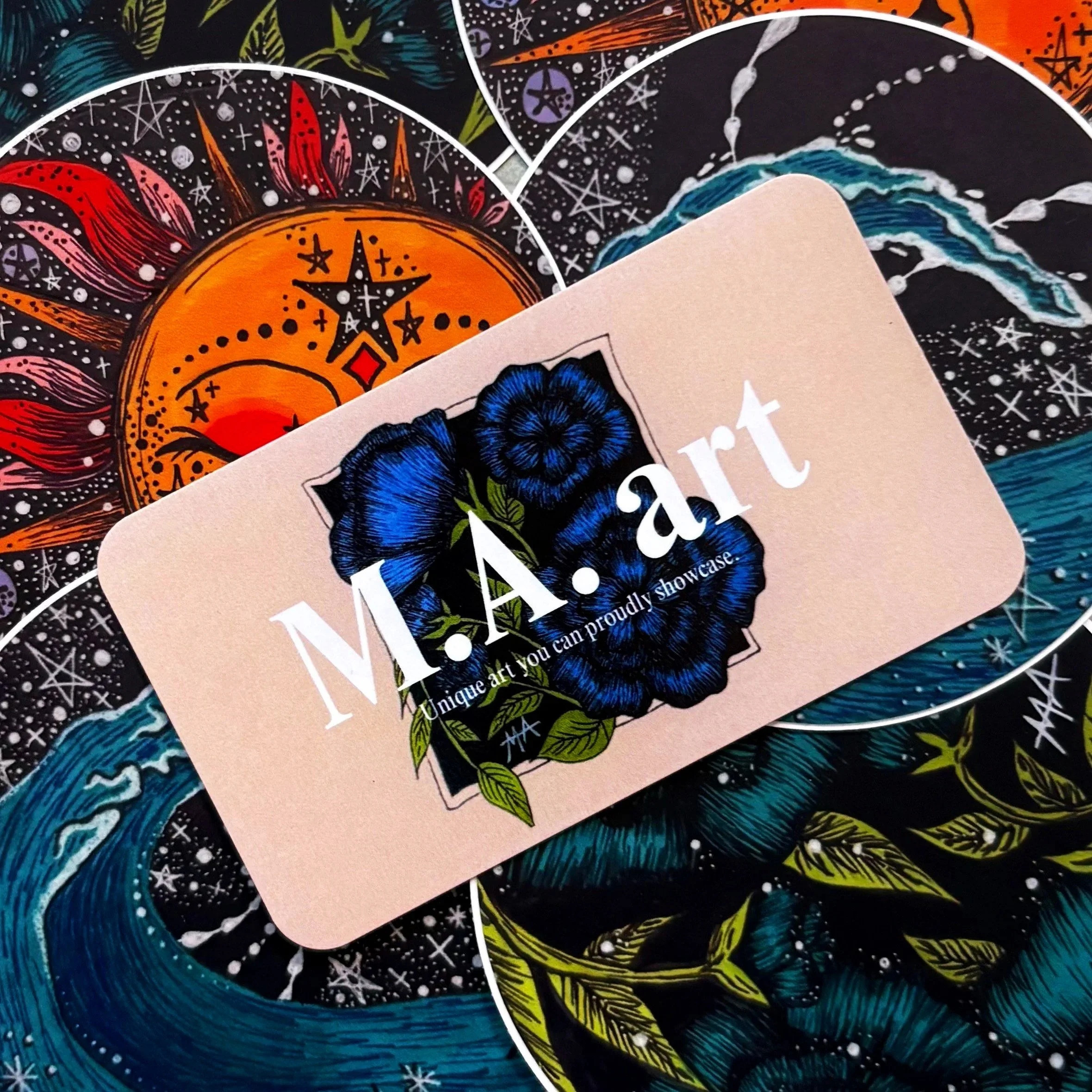 A business card with blue flowers and the words 'M.A.R.T. Unique art you can proudly showcase' placed on oracle tarot card stickers with cosmic and celestial designs.