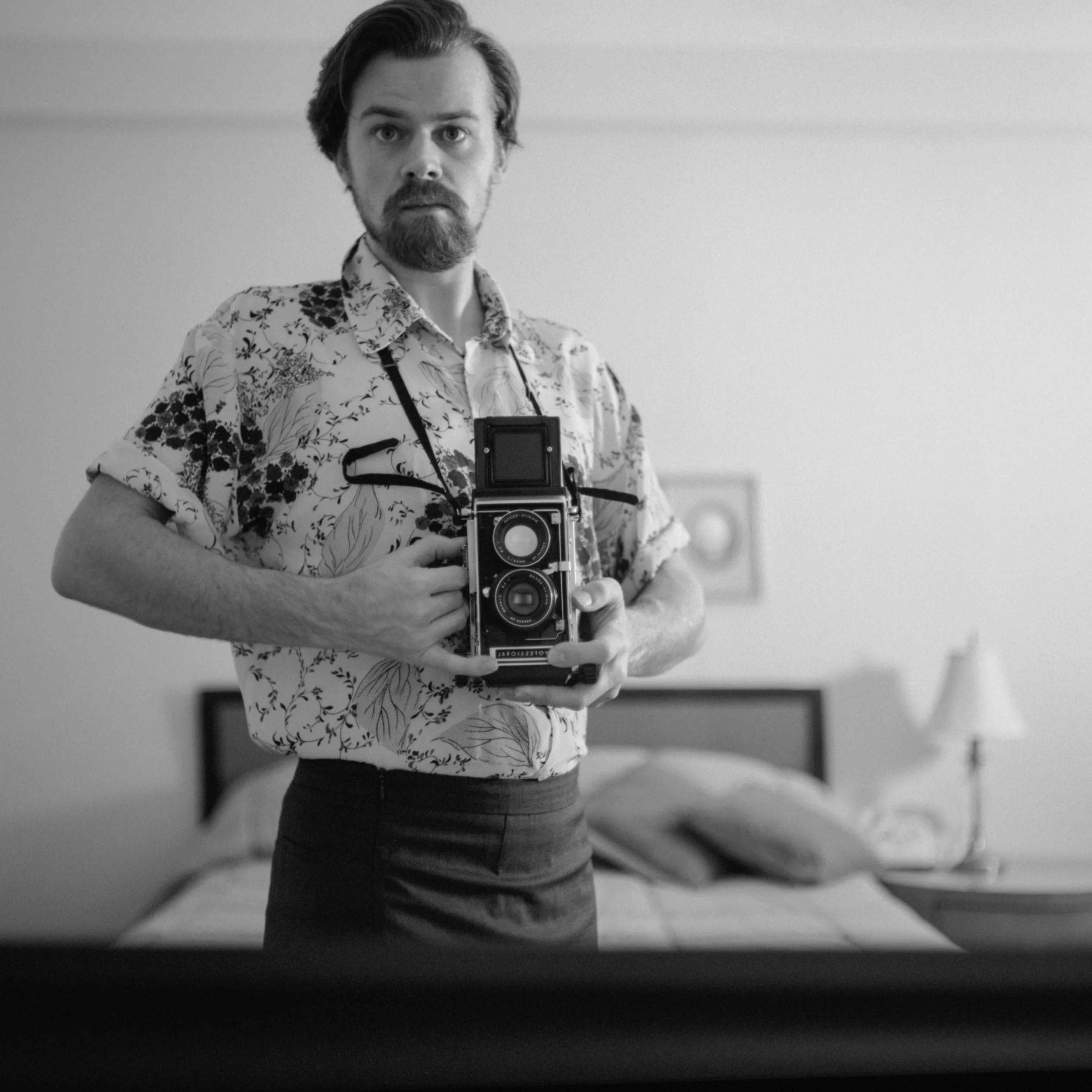 Person in floral shirt holding a vintage camera, standing in a room with a bed and lamp in the background.