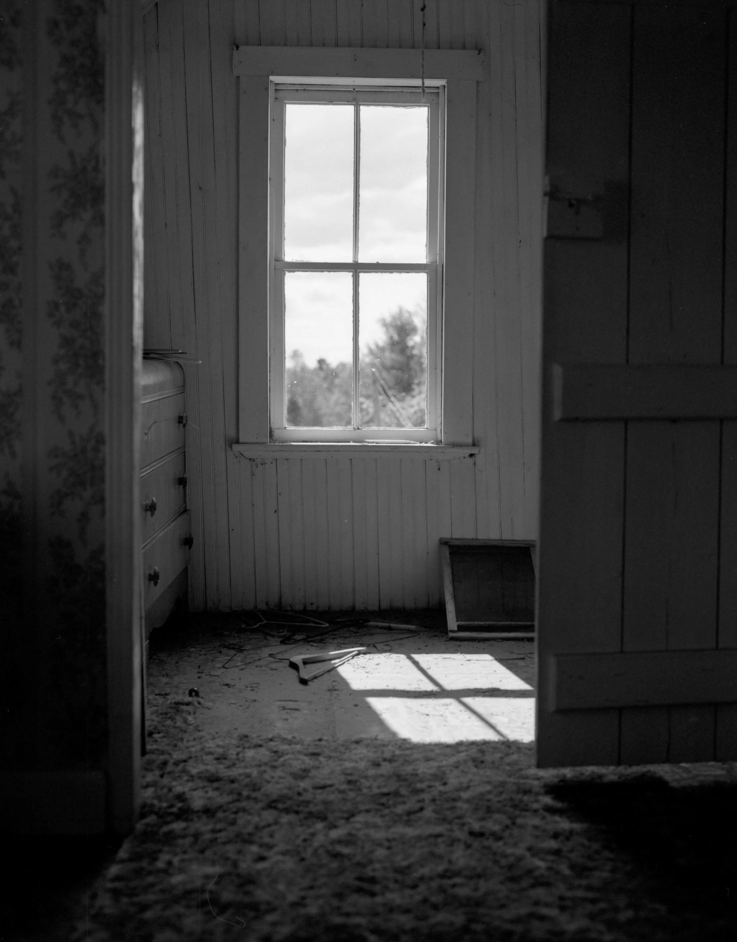 Black and white photo of an empty room with a window, showing sunlight casting a shadow on the dusty floor, and a closed door partially visible.