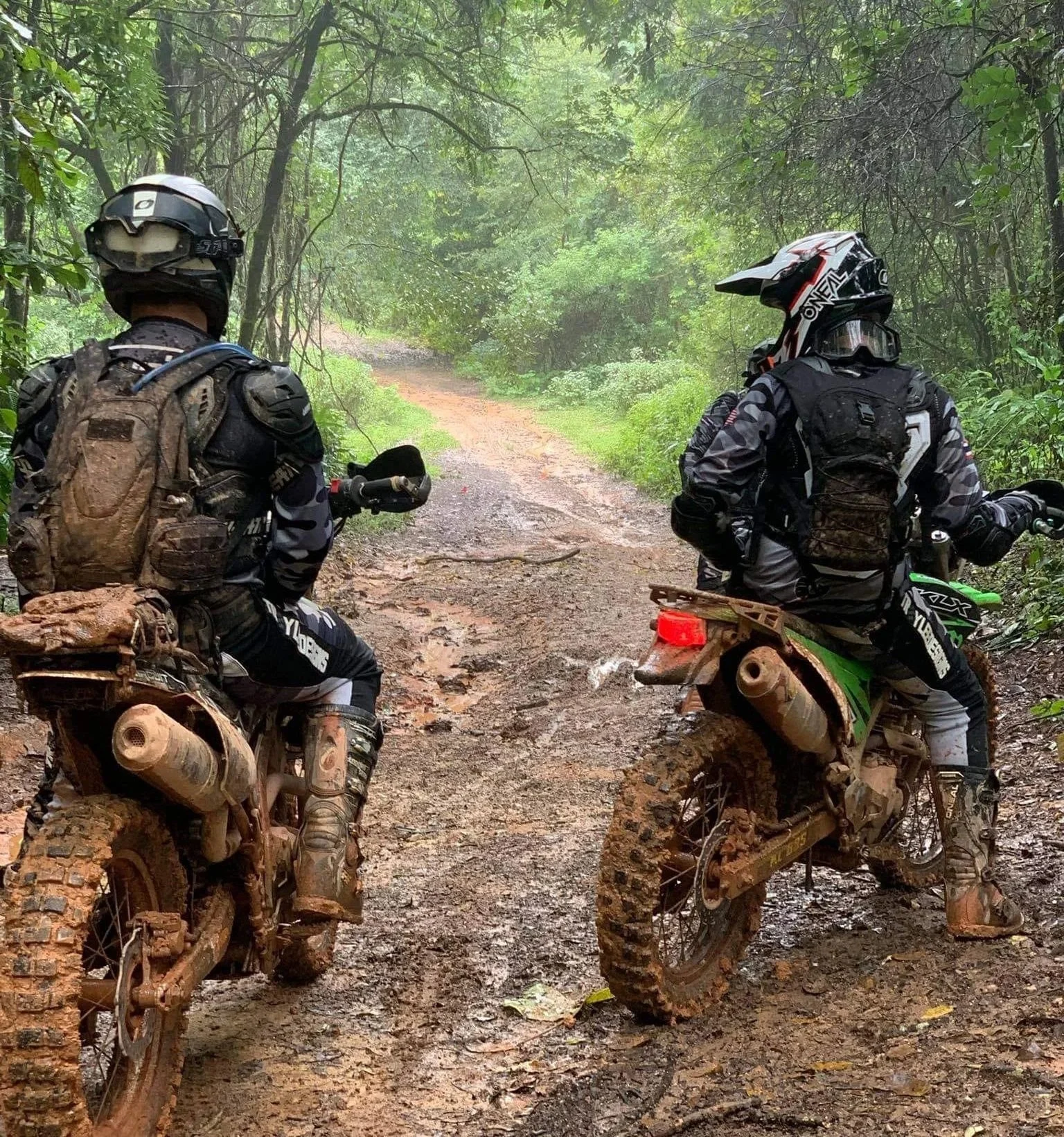 Two off-road motorcycle riders wearing helmets and gear, sitting on muddy dirt bikes on a wooded trail.