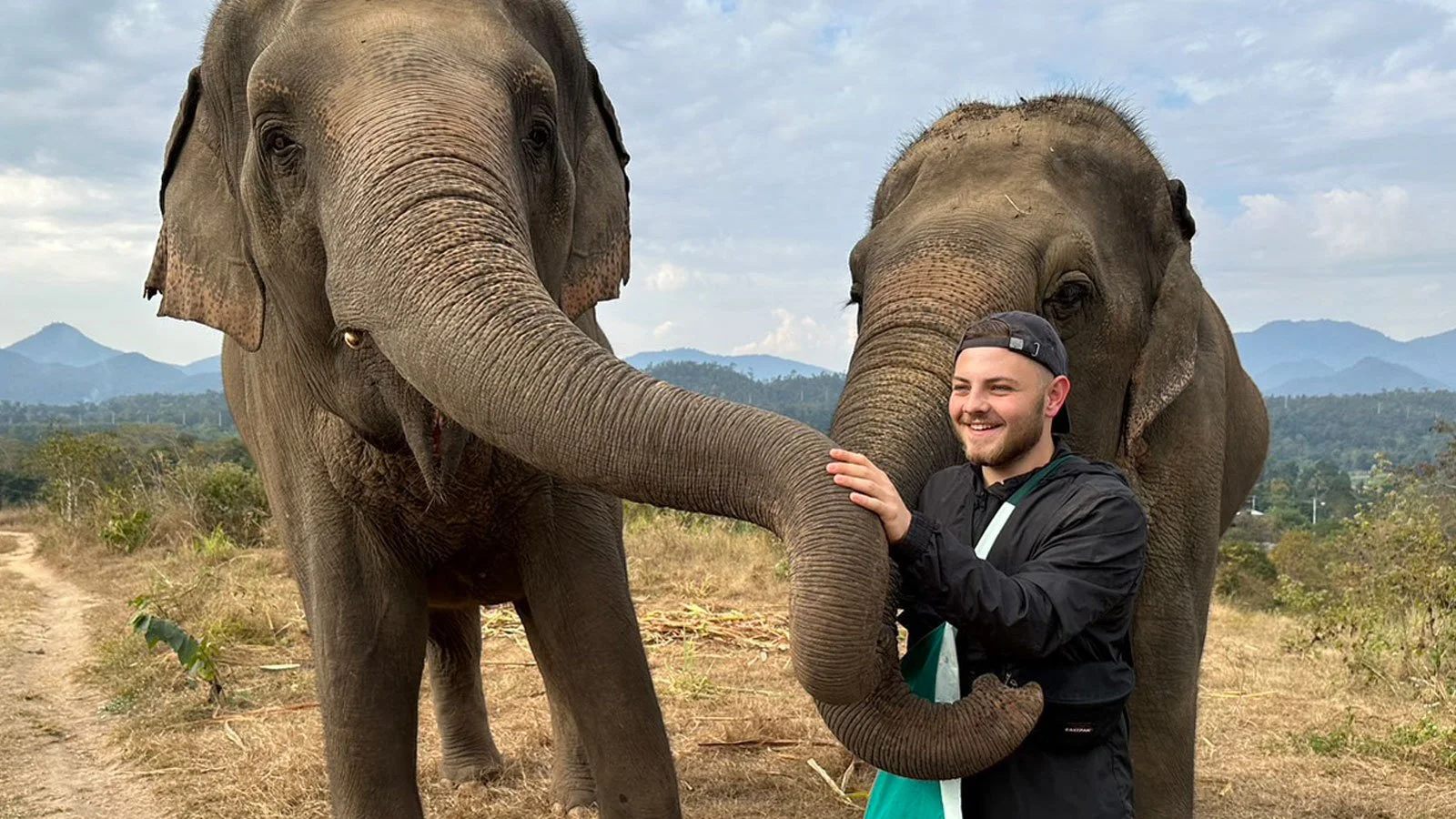 A young man in a black jacket and baseball cap smiling and petting two elephants in an open field with mountains in the background.