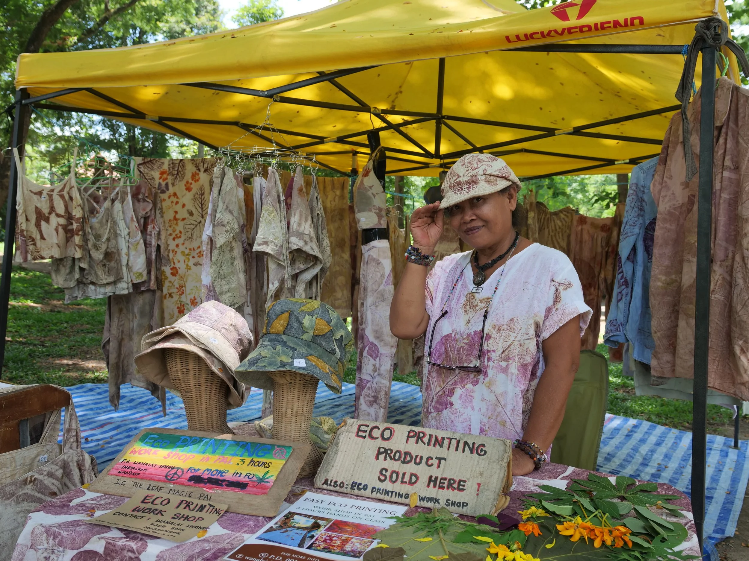 A woman standing at an outdoor market stall displaying eco-printed clothing and accessories. The stall has a yellow canopy, and various shirts, hats, and fabric pieces are hanging or displayed on tables and mannequin heads. She is wearing a white shirt with a pink pattern, a hat, and jewelry, and is adjusting her hat while smiling at the camera.