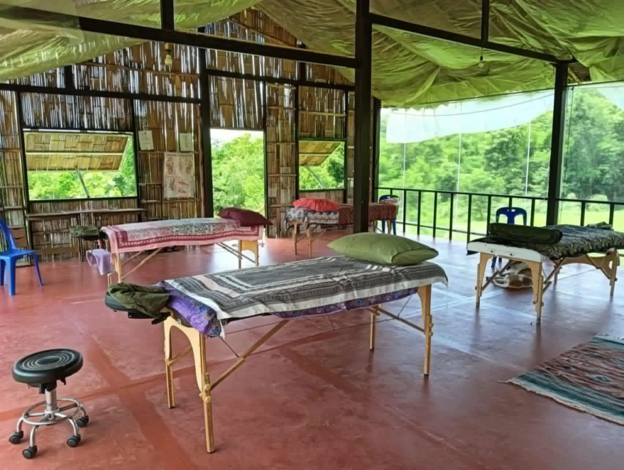 Open-air bamboo and wooden structure with several massage tables, pillows, a rolling stool, chairs, and a colorful rug, surrounded by greenery outside.