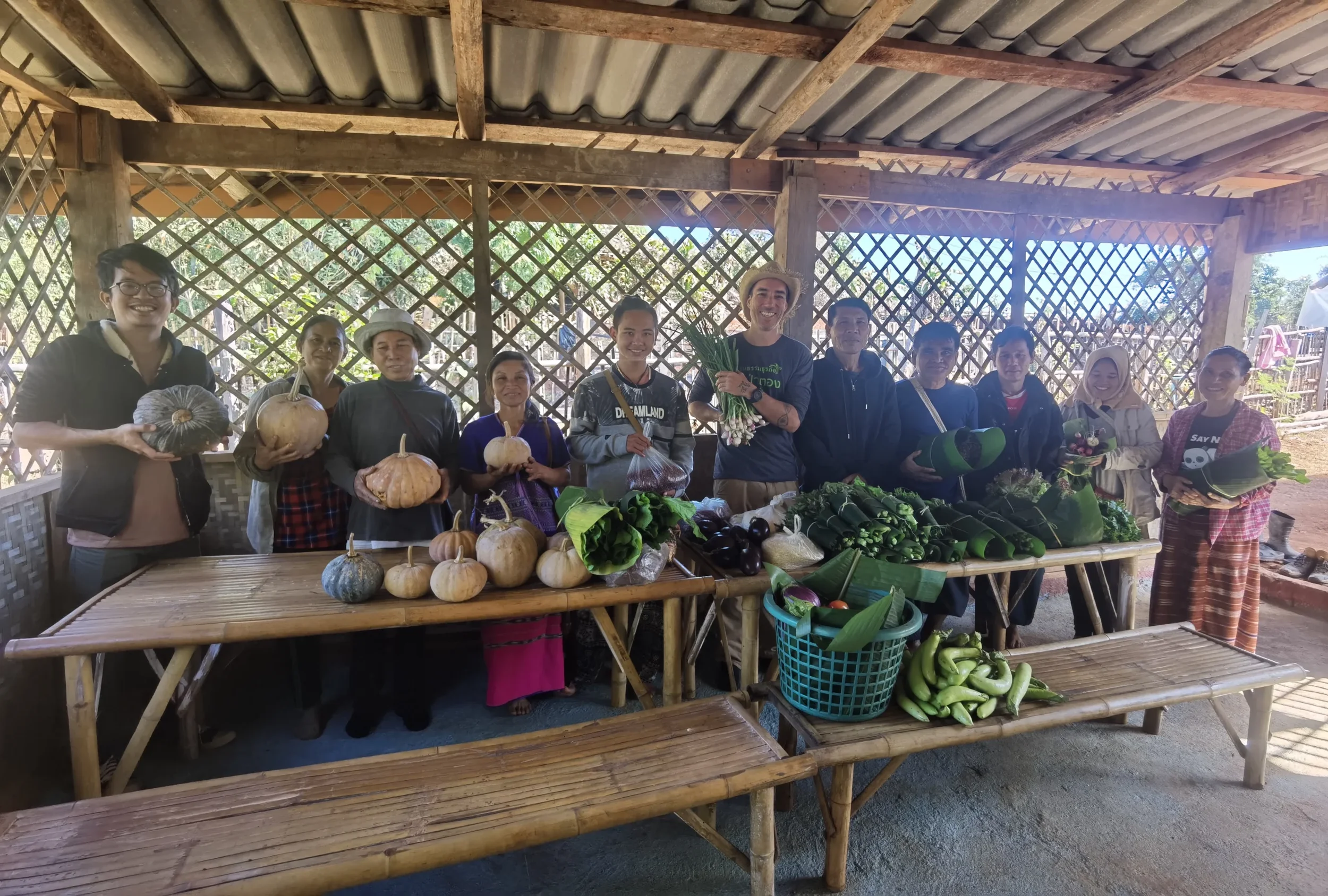Group of people standing behind a bamboo table displaying various vegetables inside a wooden shelter with lattice walls.