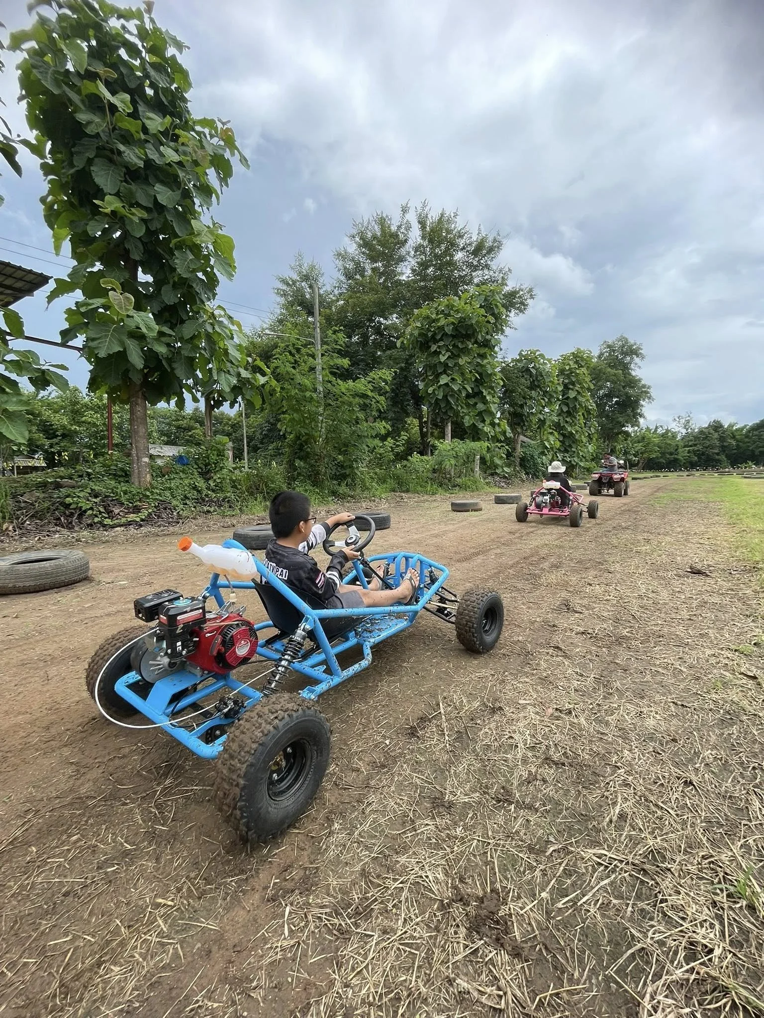 Children driving go-karts on a dirt track surrounded by green trees under a cloudy sky.