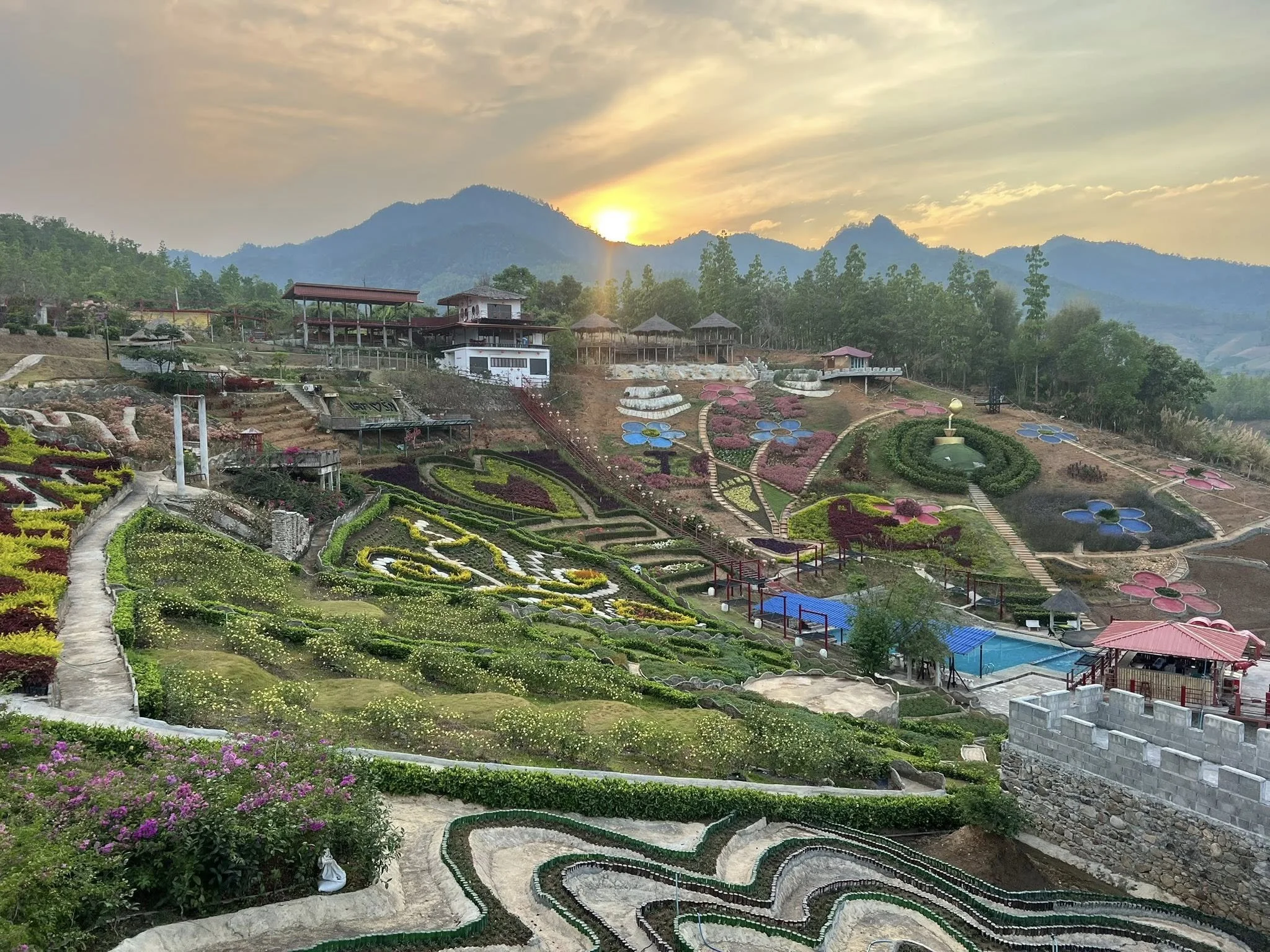 Scenic view of a colorful terraced garden with various flowers, pathways, and small structures at sunset with mountains in the background.
