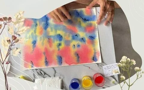 Hands placing or opening a colorful watercolor portrait or artwork on a flat surface surrounded by flowers, paints, and art supplies.