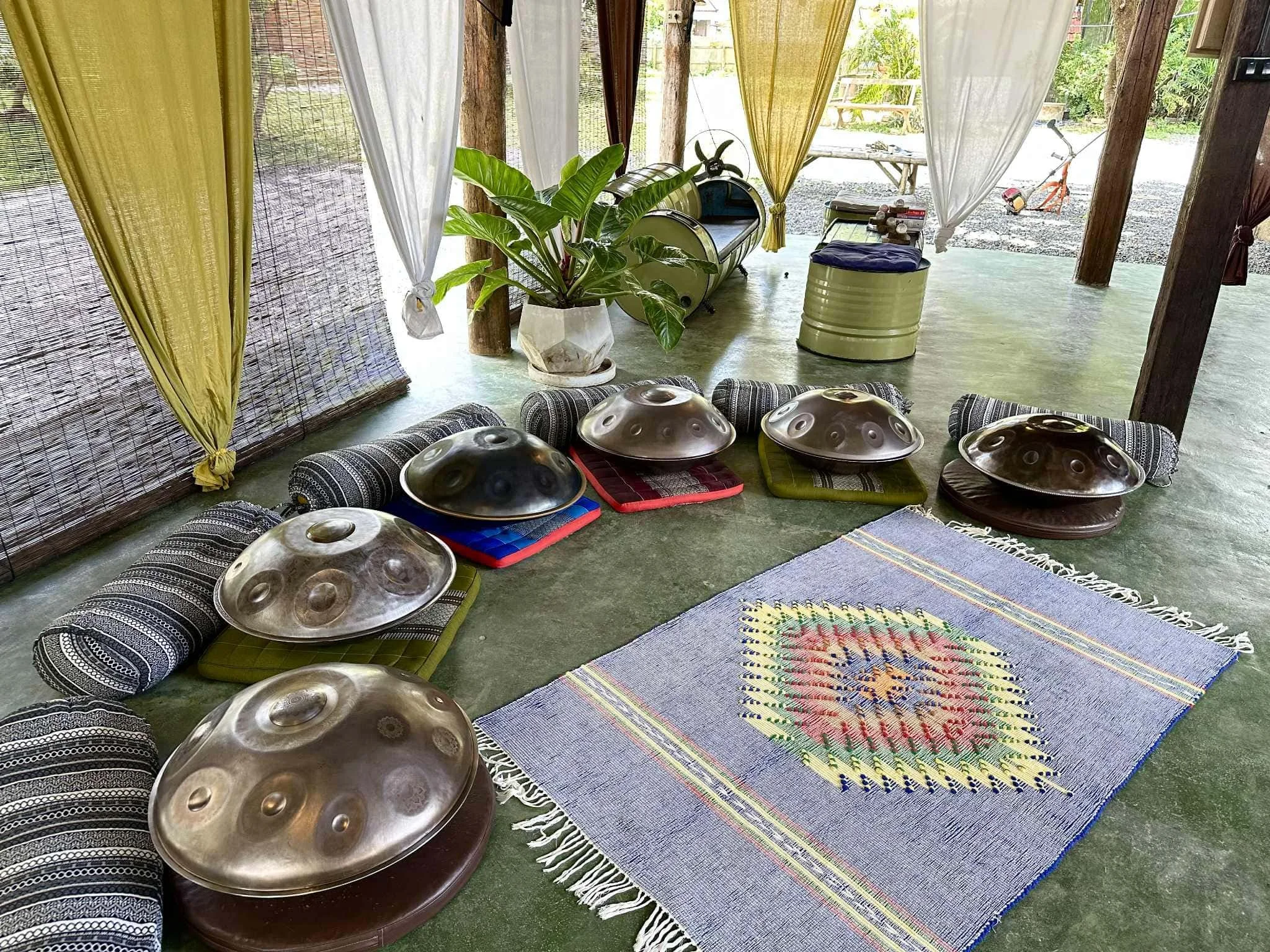 Indoor space set up for a musical or meditation session with four metal handpans on cushions and rugs, a colorful woven mat, potted plants, curtain partitions, and outdoor scenery visible through open curtains.