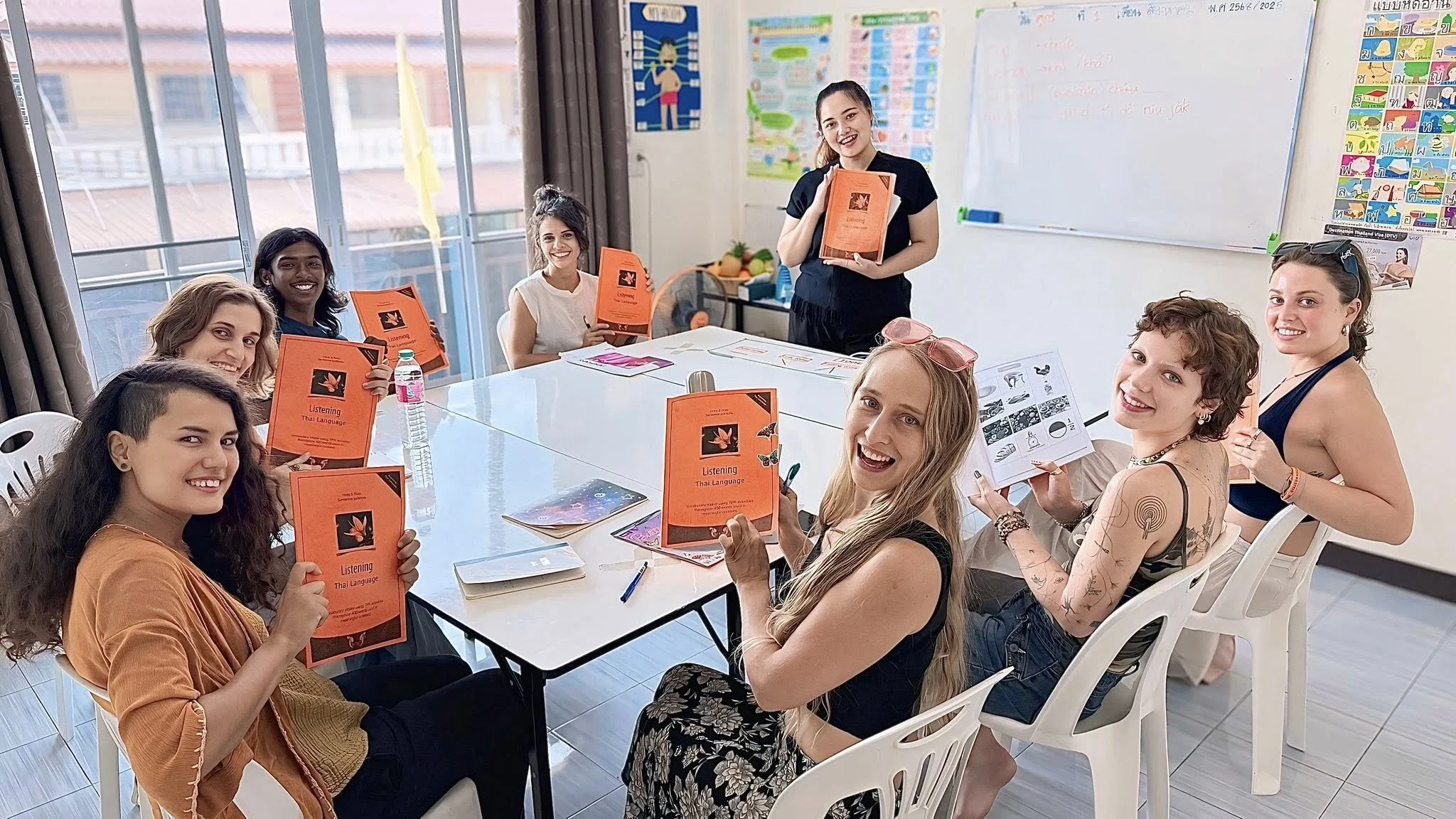 A group of women sitting around a table in a classroom, holding orange language learning booklets titled 'Listening Thai Language.' Two women are standing, conducting a class or presentation, and smiling at the camera. The classroom has a whiteboard, colorful posters, and large windows with curtains.