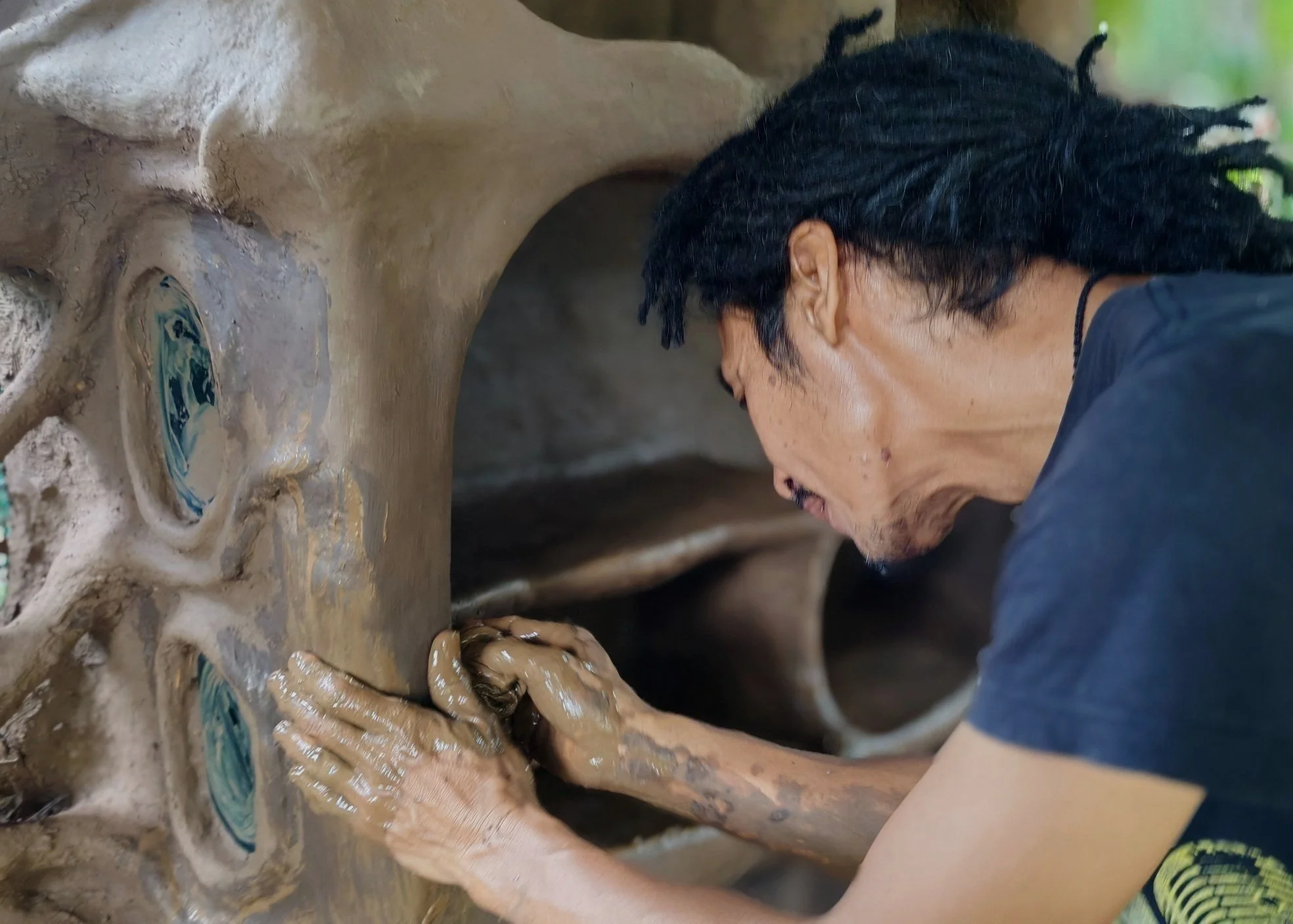 A person with dreadlocks working with clay pottery, shaping it with both hands.