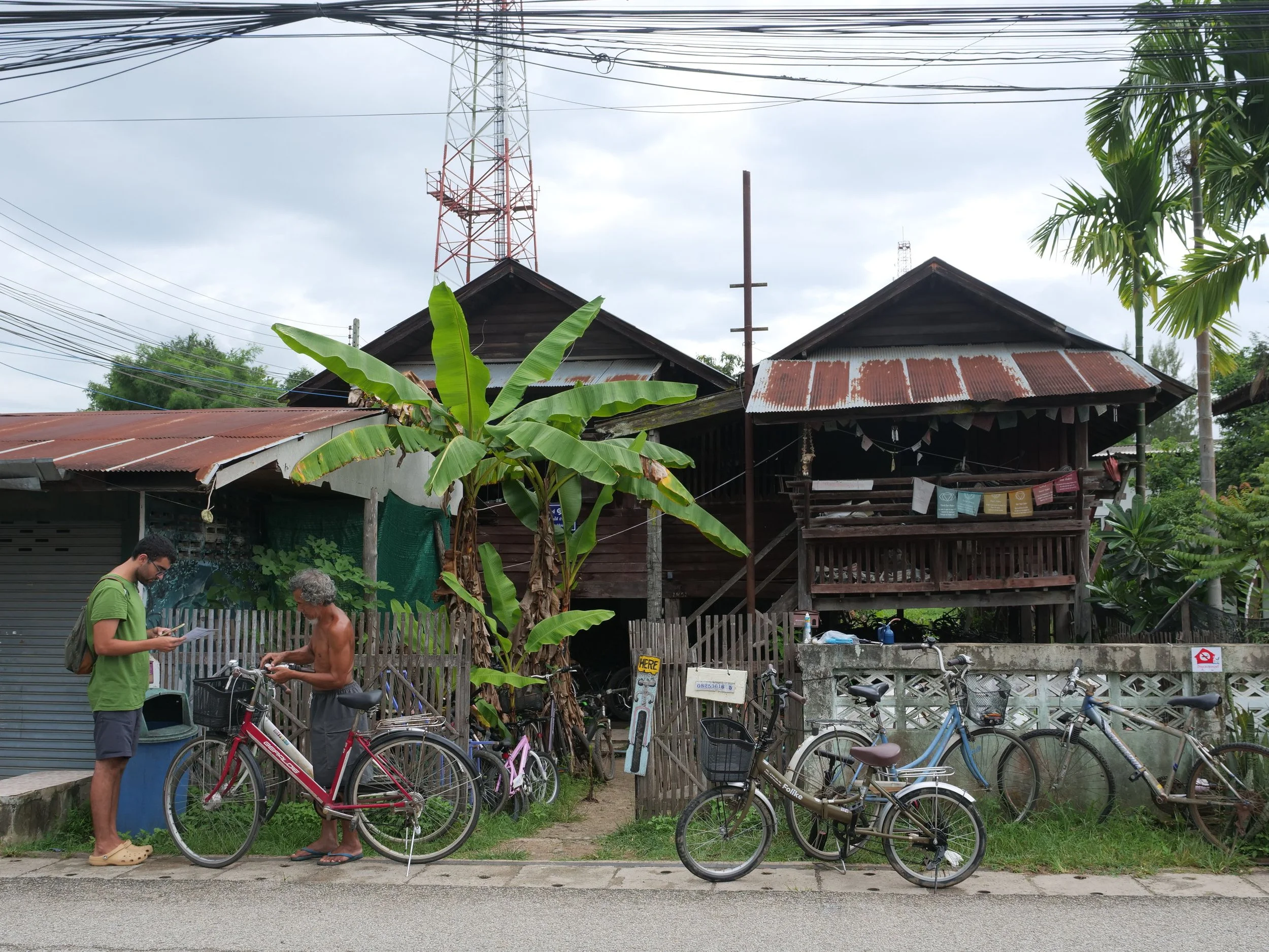 Two men with bicycles on a sidewalk near a wooden house. One man is shirtless, older with gray hair. The other man is younger, wearing glasses and a green T-shirt. The house is elevated, with a rusty metal roof, and surrounded by green plants including a banana tree. Bicycles are parked along the fence, and there is a banana tree and utility poles and power lines in the background.