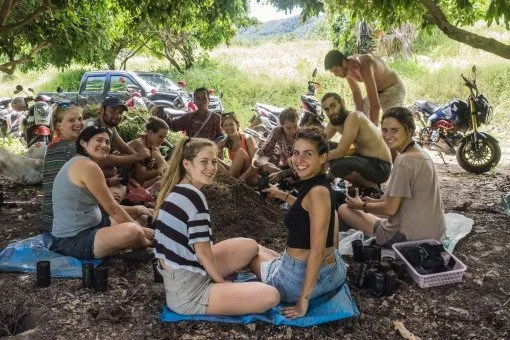 Group of friends sitting on the ground outdoors under a tree, having a picnic with motorcycles and cars in the background.