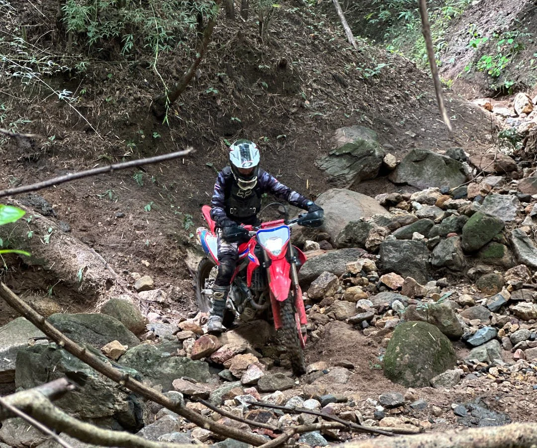 A person riding a red dirt bike through a rocky forest trail wearing a helmet and protective gear.