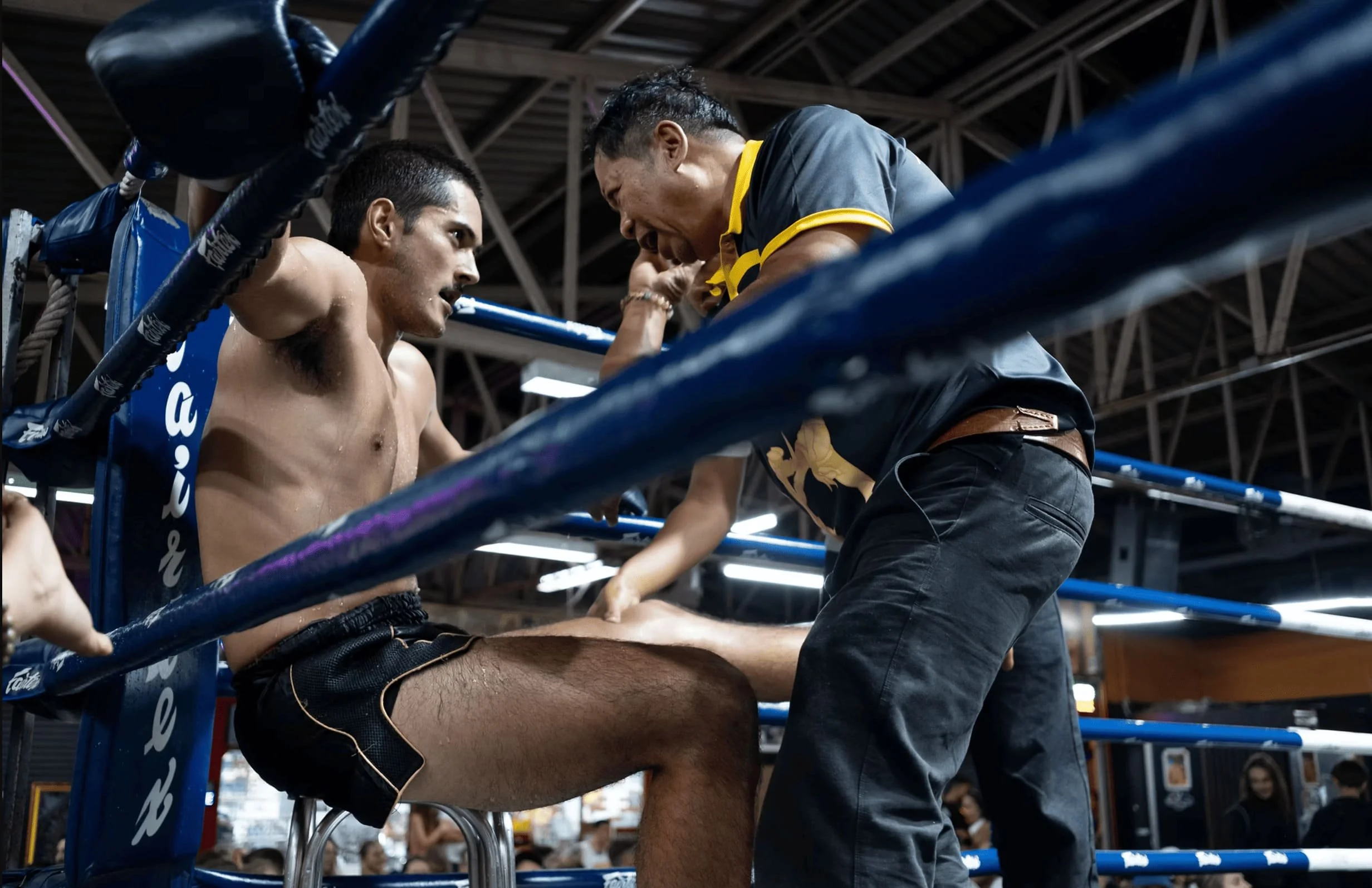 A boxer sitting on a stool receiving coaching and encouragement from his trainer inside a boxing ring.
