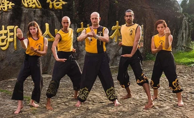 Five martial artists in yellow and black uniforms practicing martial arts outside on a stone ground with a large rock and Chinese characters in the background.