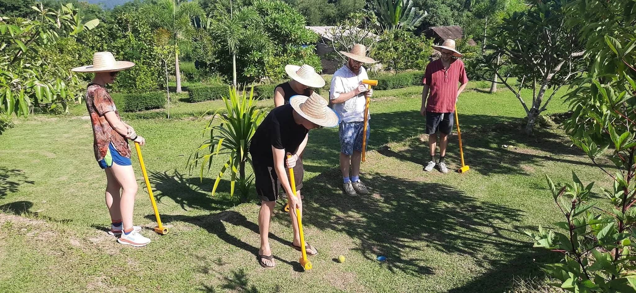 Group of five people wearing sun hats playing croquet outdoors in a lush garden with trees and bushes.