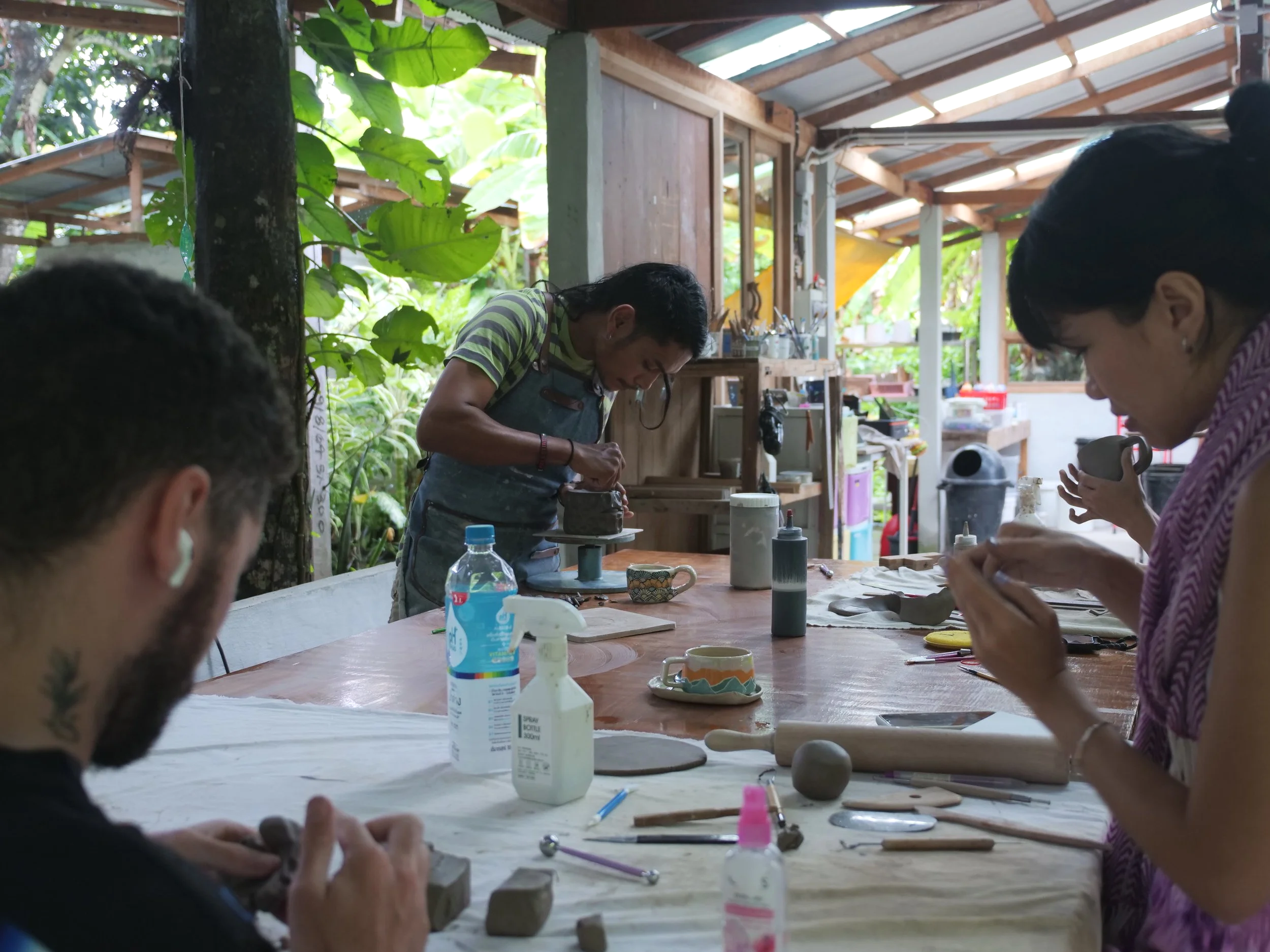 People working on clay pottery in a rustic, open-air workshop with a wooden table and lush greenery outside.