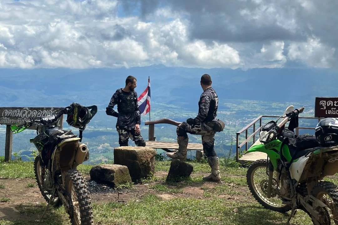 Two motorcyclists in riding gear with backpacks are resting on a hilltop with the scenic view of a valley and mountains in the background. They are standing near a bench, with two off-road motorcycles parked nearby.