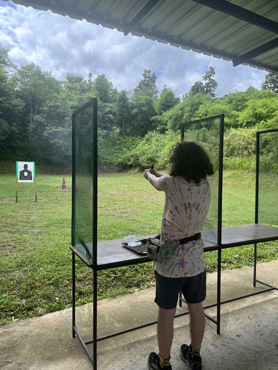 Person at a shooting range aiming a handgun at a target in the distance, with an outdoor grassy area and trees behind.
