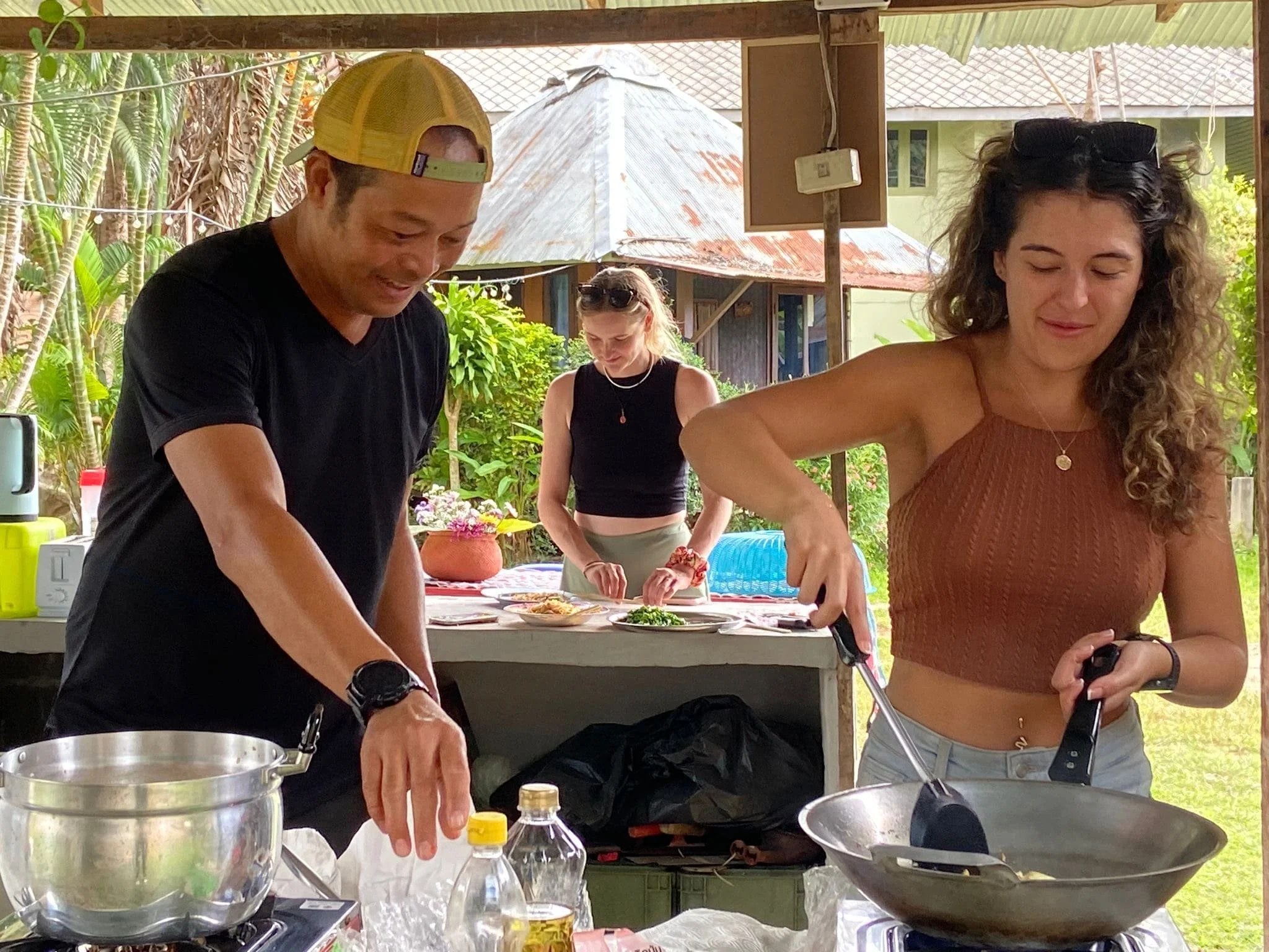 Two people cooking outdoors at a barbecue, with a woman in a brown sleeveless top and a man in a black t-shirt and yellow cap, and another woman in the background preparing food.