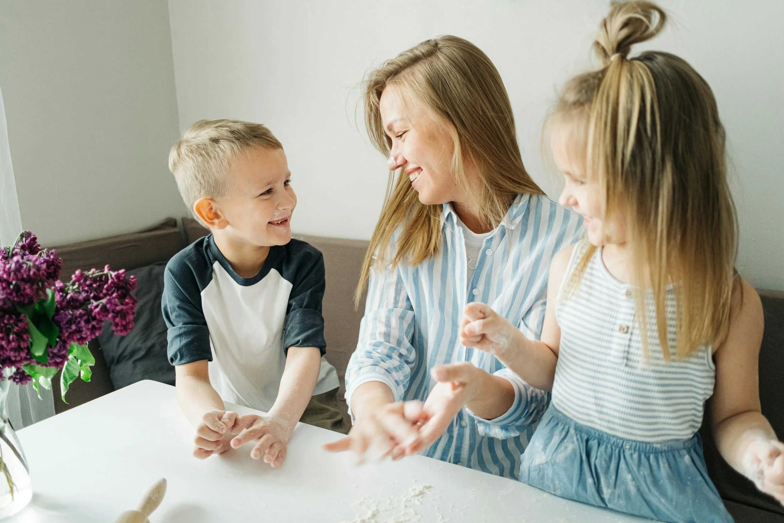 A woman and two young girls baking together in a kitchen, smiling and laughing.