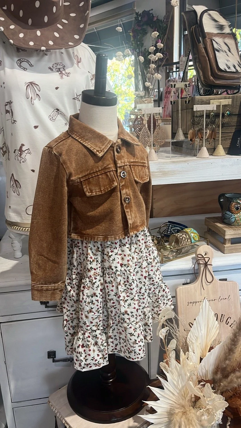 Mannequin dressed in a brown cropped denim jacket over a floral skirt, displayed in a boutique with accessories and home decor around.