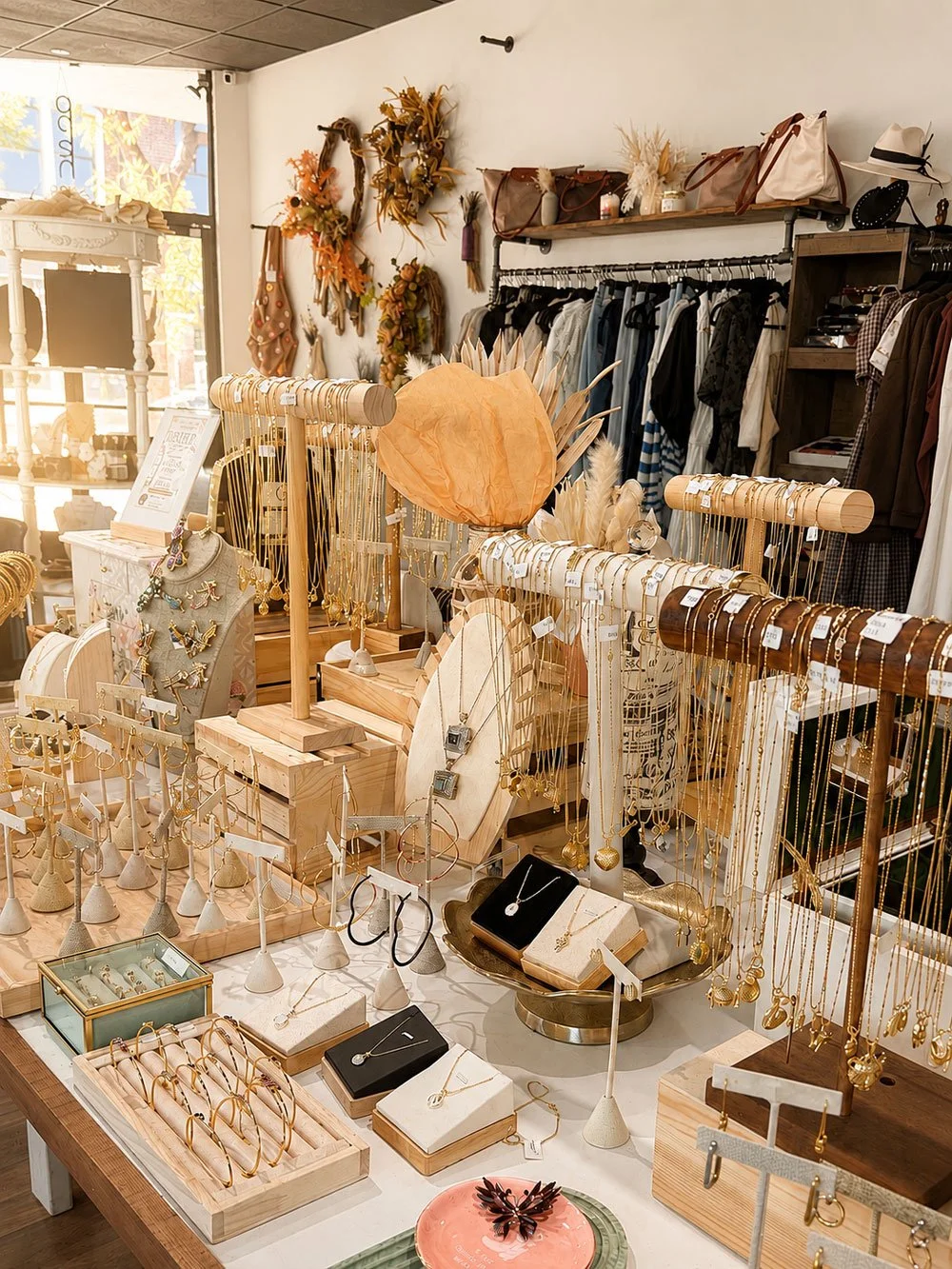 Display of jewelry including necklaces, earrings, and rings, in a boutique store with clothing and decorative wreaths in the background.