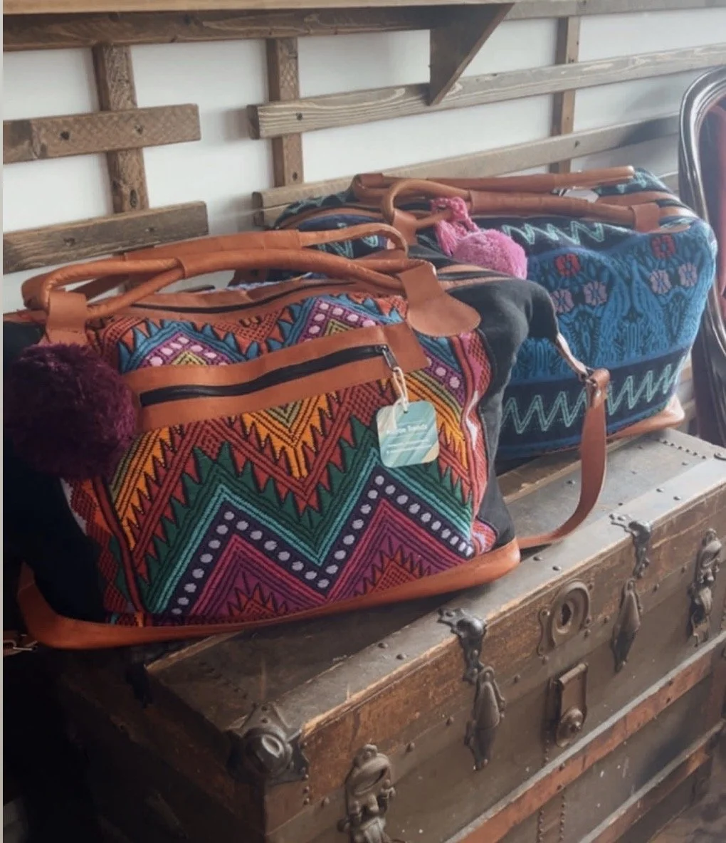 Two colorful patterned duffel bags with leather handles and pom-poms on a jewelry chest display on a rustic wooden shelf.