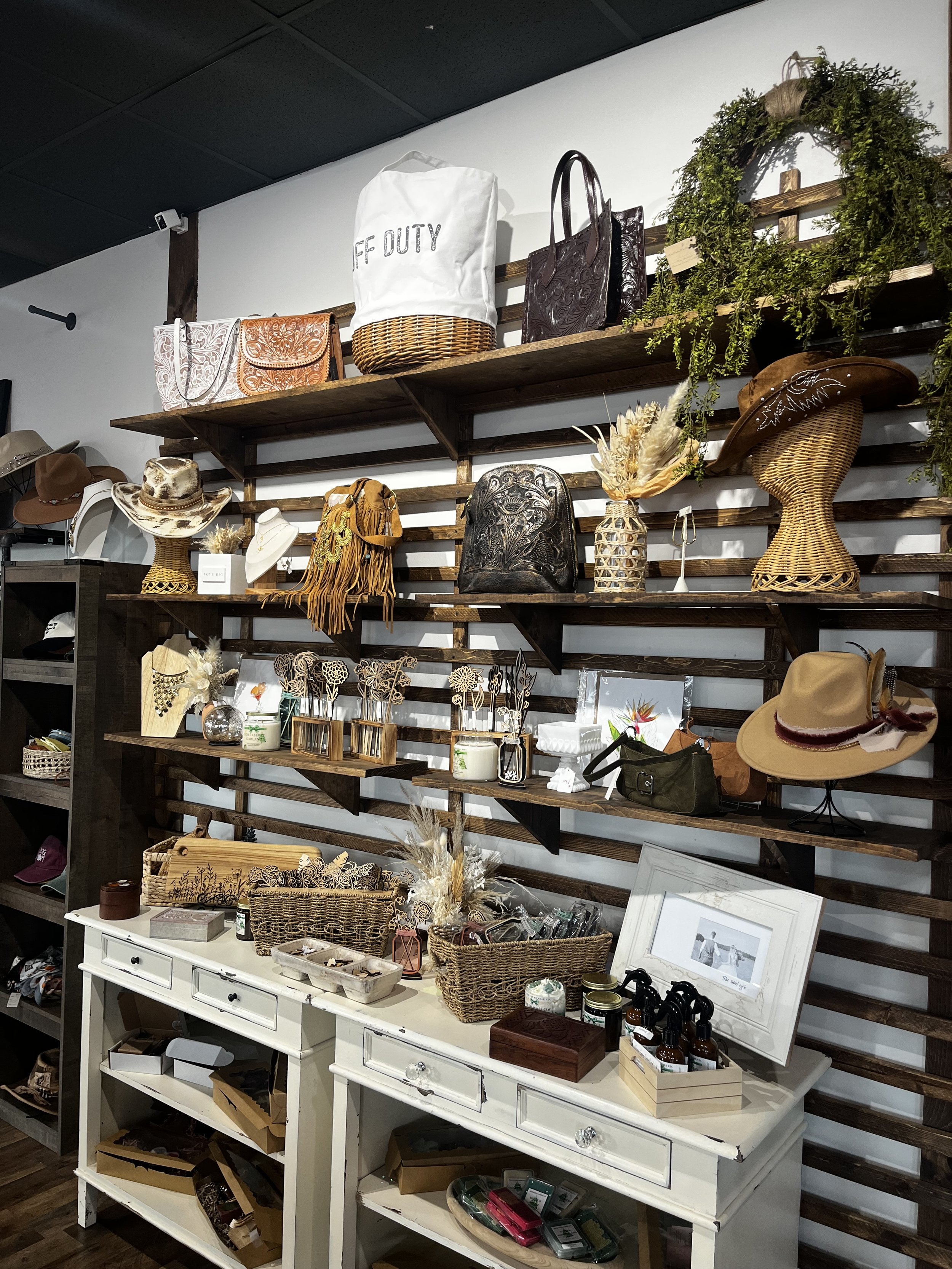Display of various fashion and home decor items including handbags, hats, picture frames, and decorative objects arranged on wooden shelves and a white table in a boutique store.