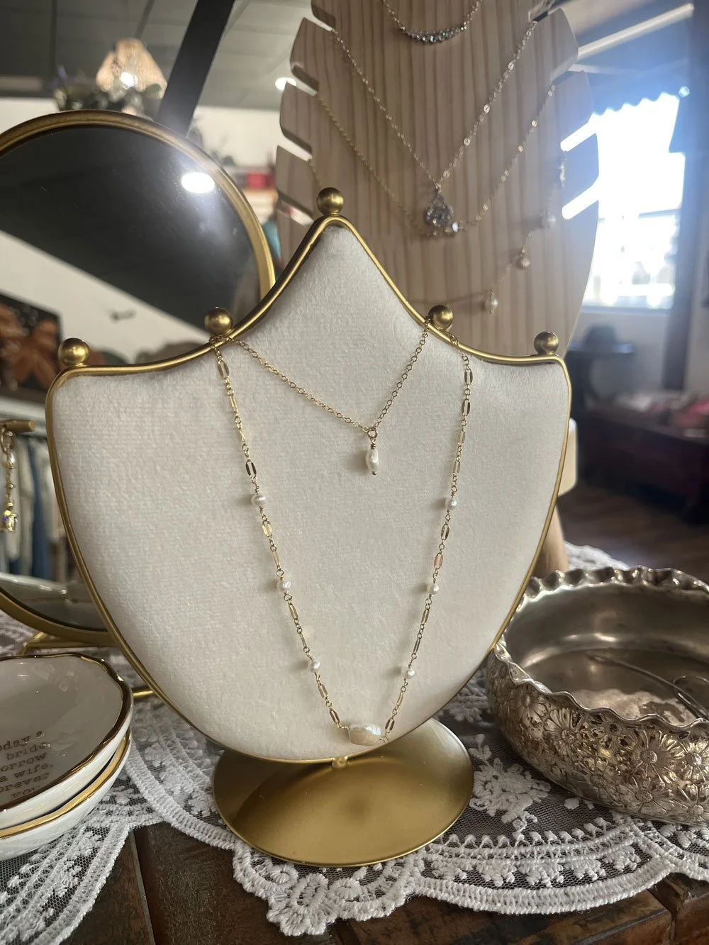 Display of gold necklaces with pearls, arranged on a cream jewelry stand with a gold outline, set on a white lace cloth. A mirror and a silver decorative bowl are also visible nearby.