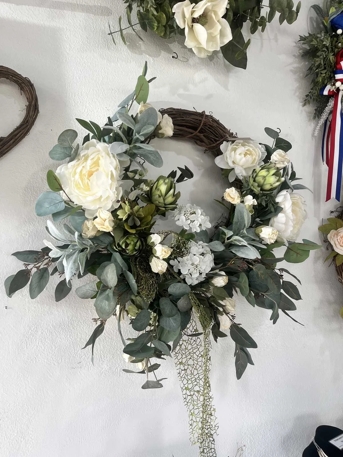 A decorative floral wreath with white roses, green and gray foliage, and artichokes, hanging on a white wall.