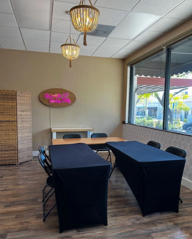 A room with wooden floors, two round chandeliers, a neon pink sign on the wall that says 'You're Like Really Pretty,' and a window showing an outdoor area with trees and buildings. There are two tables covered with black cloths and several chairs around them.