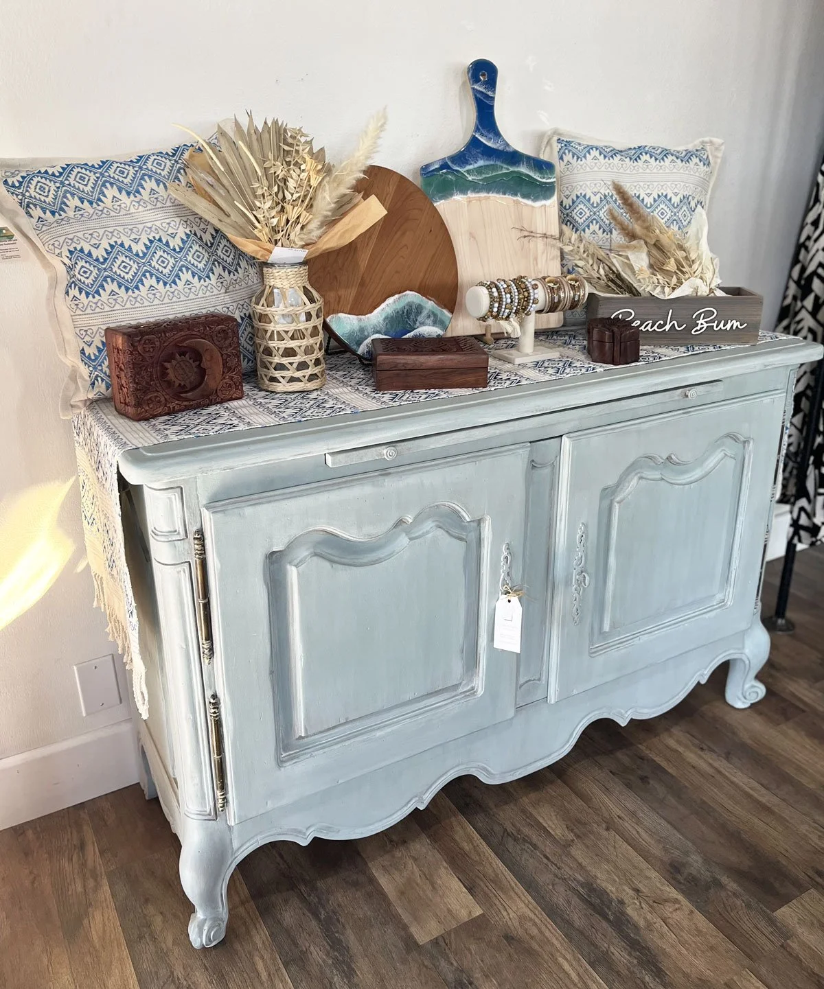 A vintage light blue sideboard decorated with tableware, a bouquet of dried flowers, wooden boxes, and jewelry display, against a white wall and wooden floor in Upland, CA.