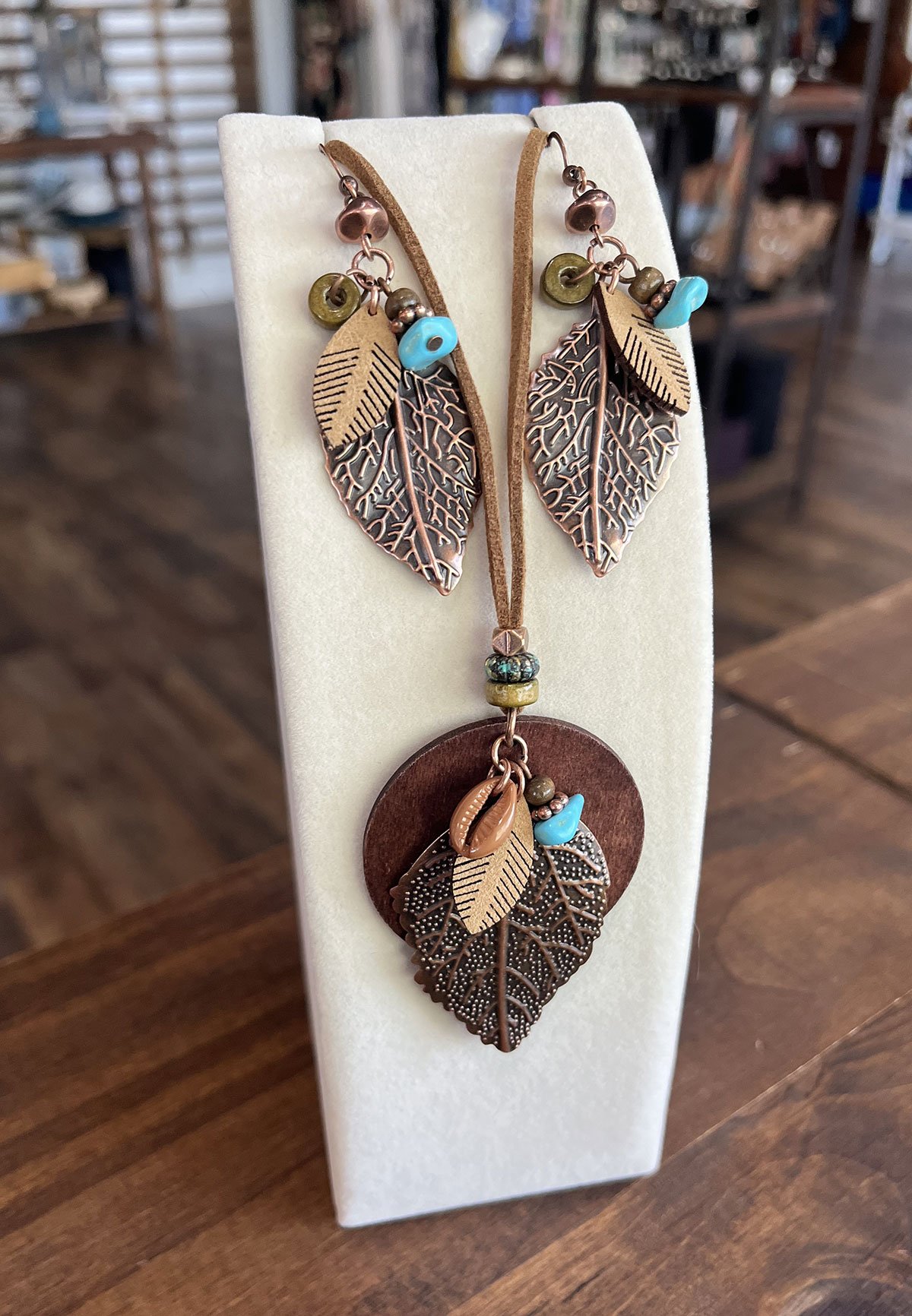 A jewelry set with leaf and bead charms on a display stand, including earrings and a necklace with a round wooden pendant, displayed on a wooden surface in Upland, CA.