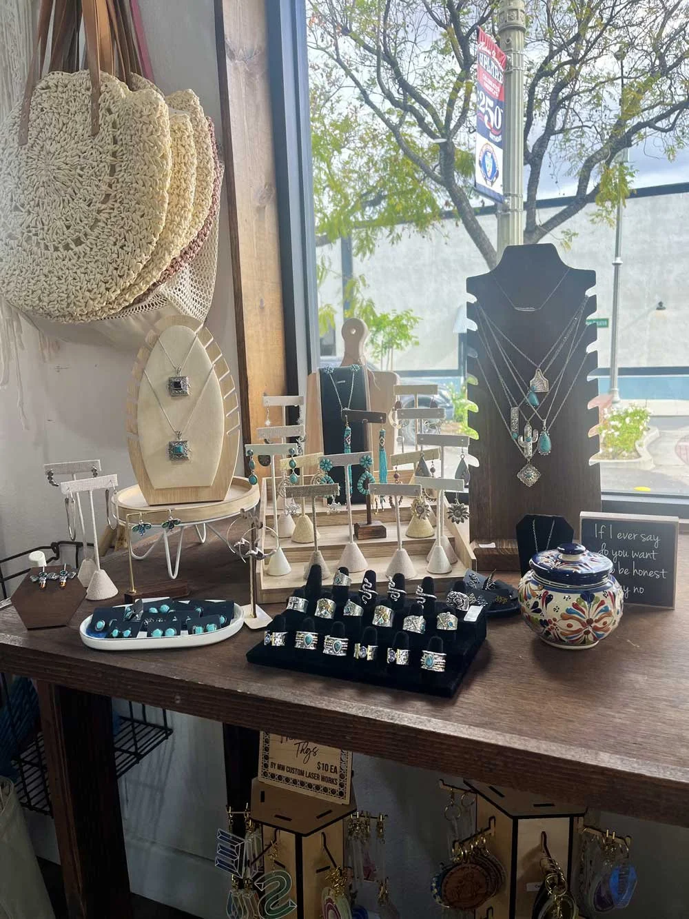 Display of jewelry including necklaces, rings, and earrings, with hats hanging on the wall in a shop window, outside trees, and a street sign visible through the window.
