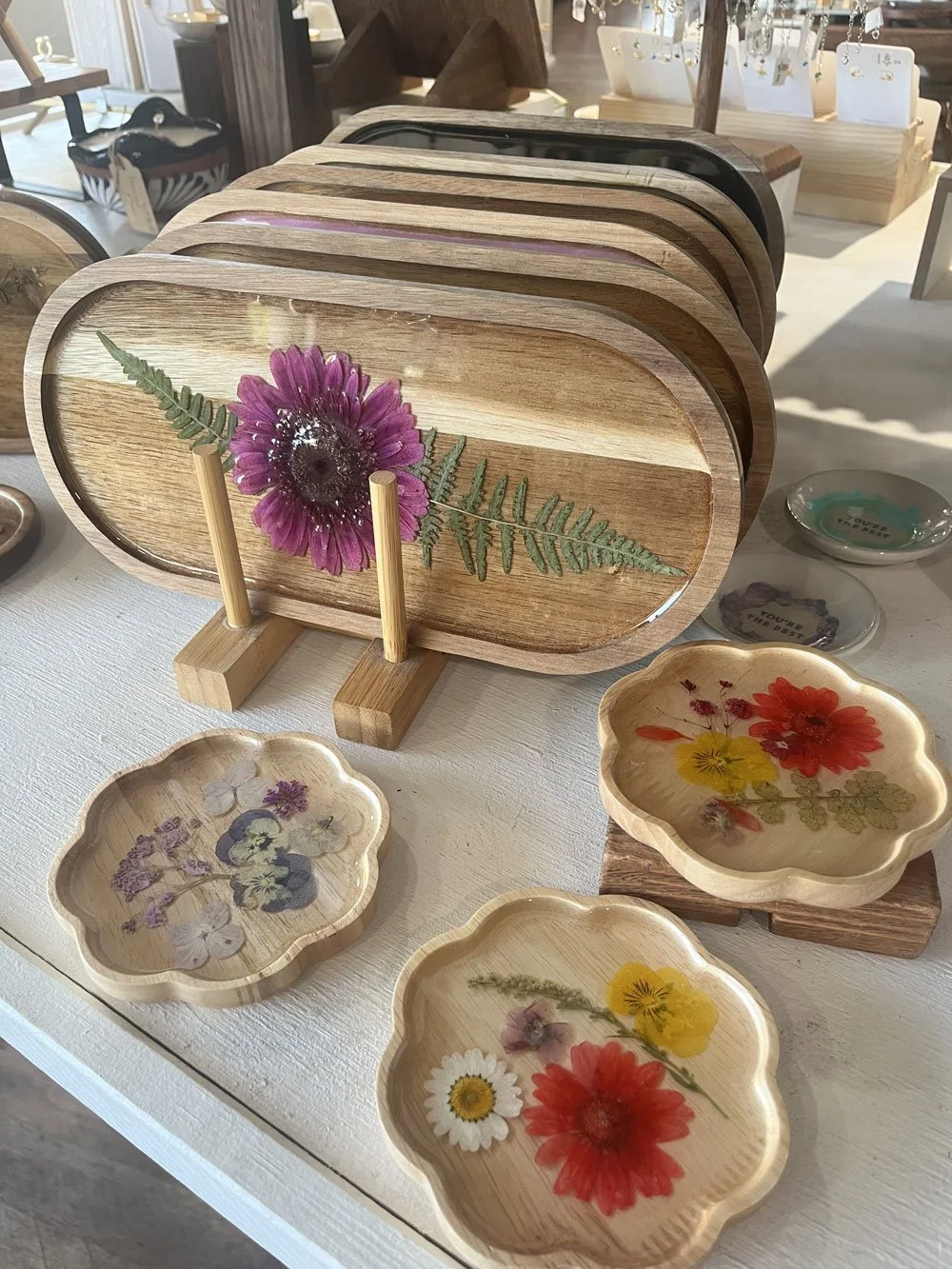 Wooden trays and bowls decorated with pressed flowers and green foliage, displayed on a white table in a retail store setting.