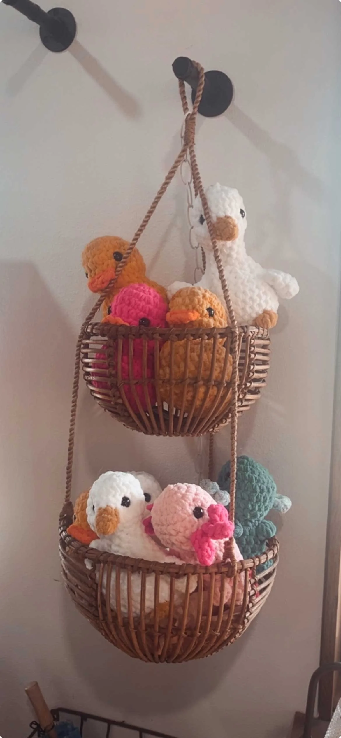 Three-tier hanging basket filled with colorful crocheted stuffed animals resembling birds and chicks, attached to a white wall.