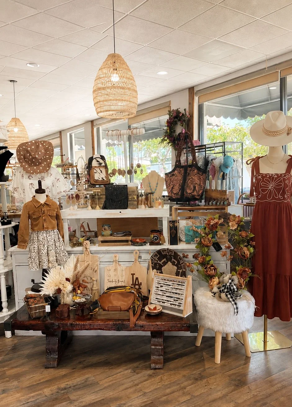 Display of women's fashion accessories and clothing, including hats, handbags, jewelry, and a dress, in a boutique store with large windows and natural lighting.