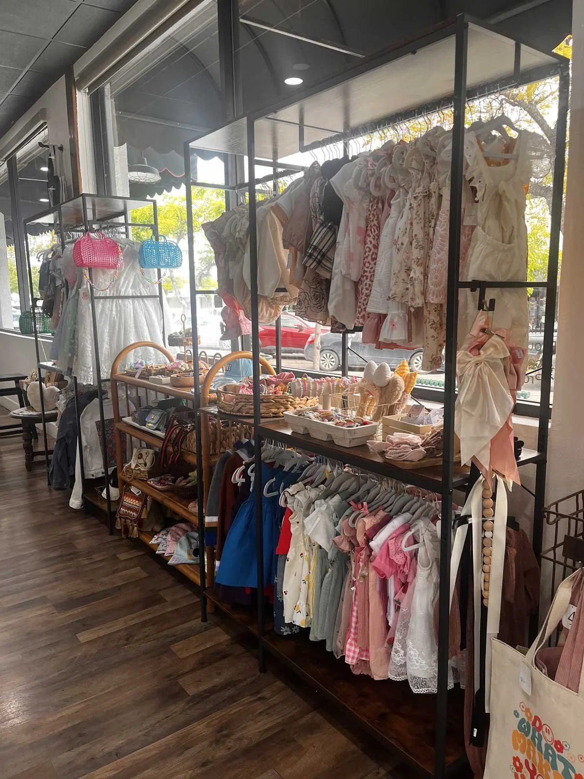 A display of children's clothing, accessories, and toys in a store. Toddler dresses, hats, and small outfits are hanging on racks and shelves, with some pastel-colored items and decorative baskets on the tables.