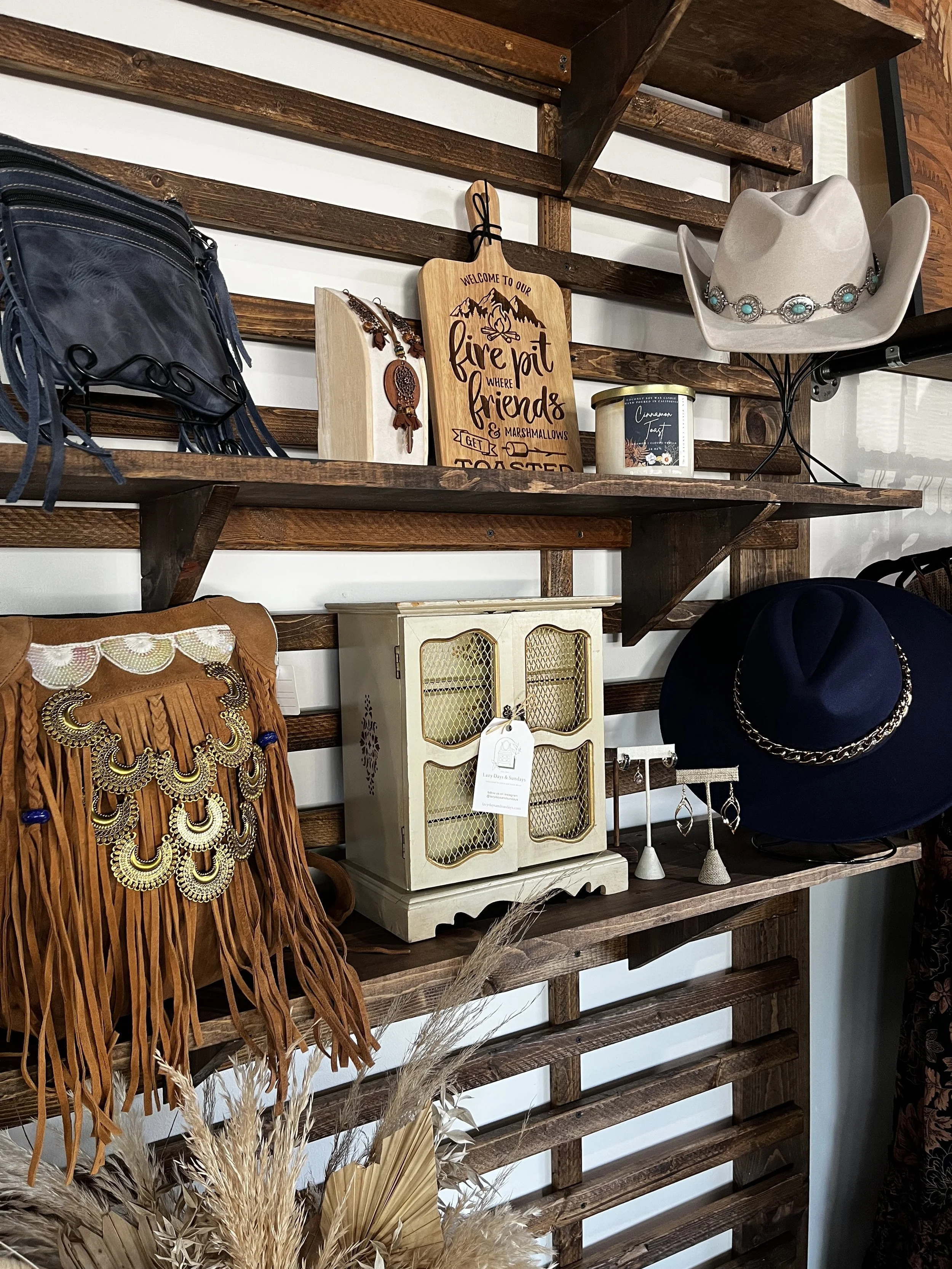 Rustic boutique shelf display featuring a leather fringe handbag with gold accents, engraved wooden cutting board sign, turquoise concho cowboy hat, jewelry, and home décor pieces. Available at Rustic and Modern Collective in Upland, CA.