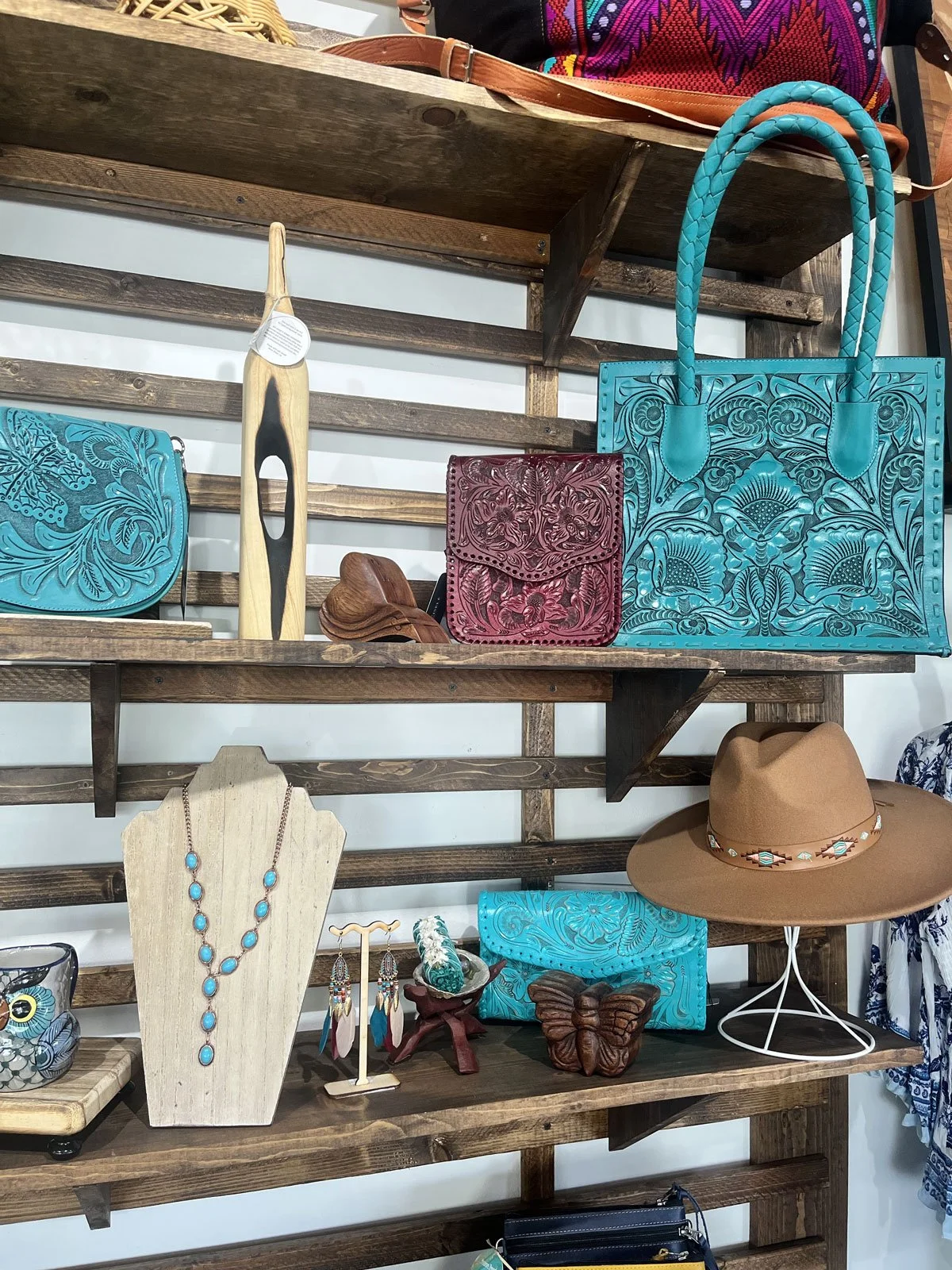 Display of colorful handbags, jewelry, and accessories on wooden shelves, including a turquoise leather bag, a red embossed leather wallet, and a large turquoise tote with floral embroidery.