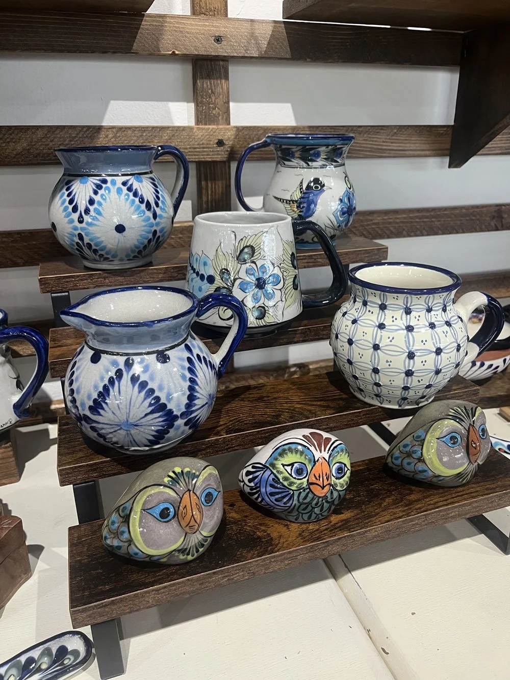 Ceramic and painted stones displayed on a wooden shelf, including painted owl stones and ceramic mugs and pitchers with blue and white patterns.