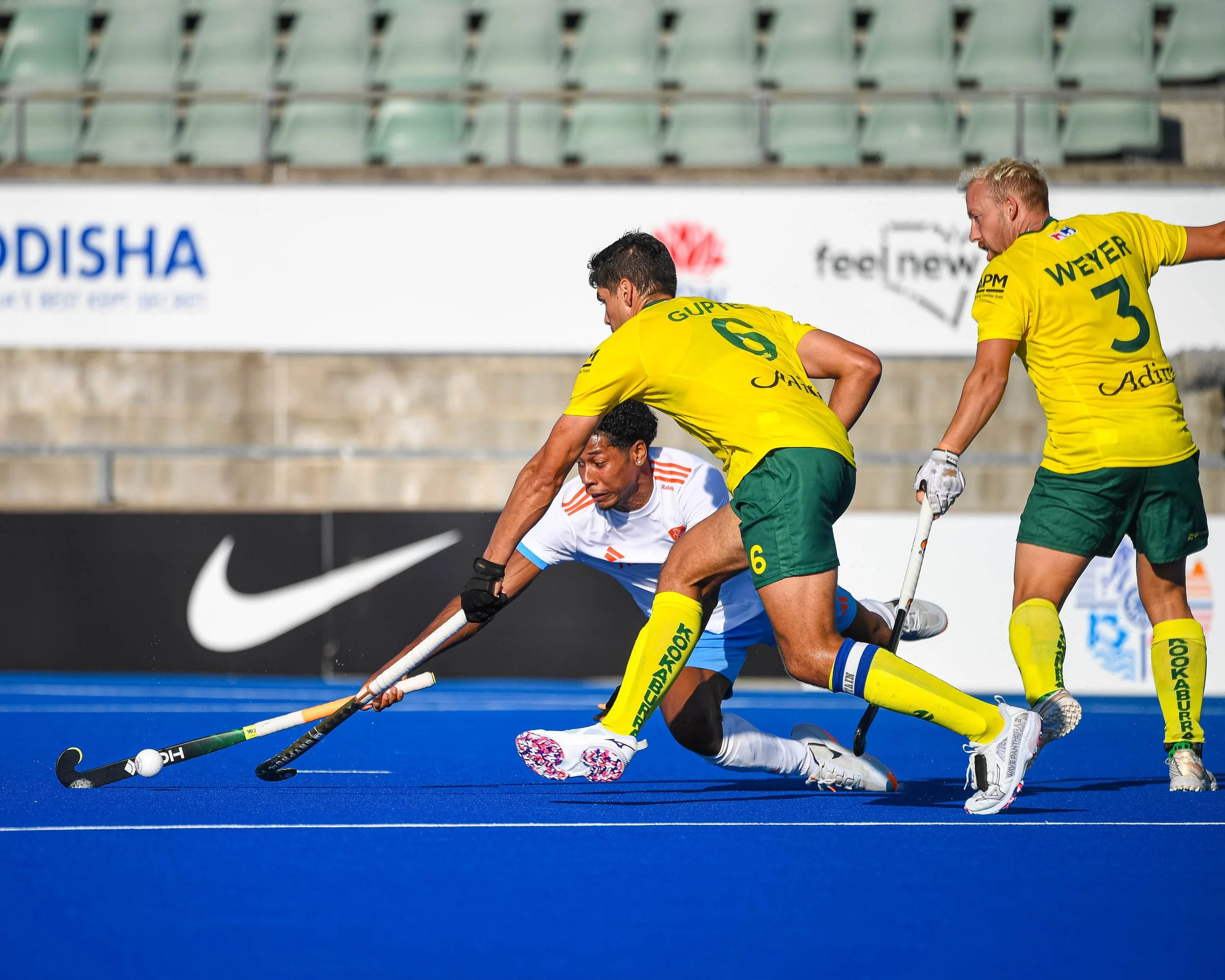 Two field hockey players in yellow uniforms and a player in a white uniform competing for control of a ball on a blue turf field during a match.