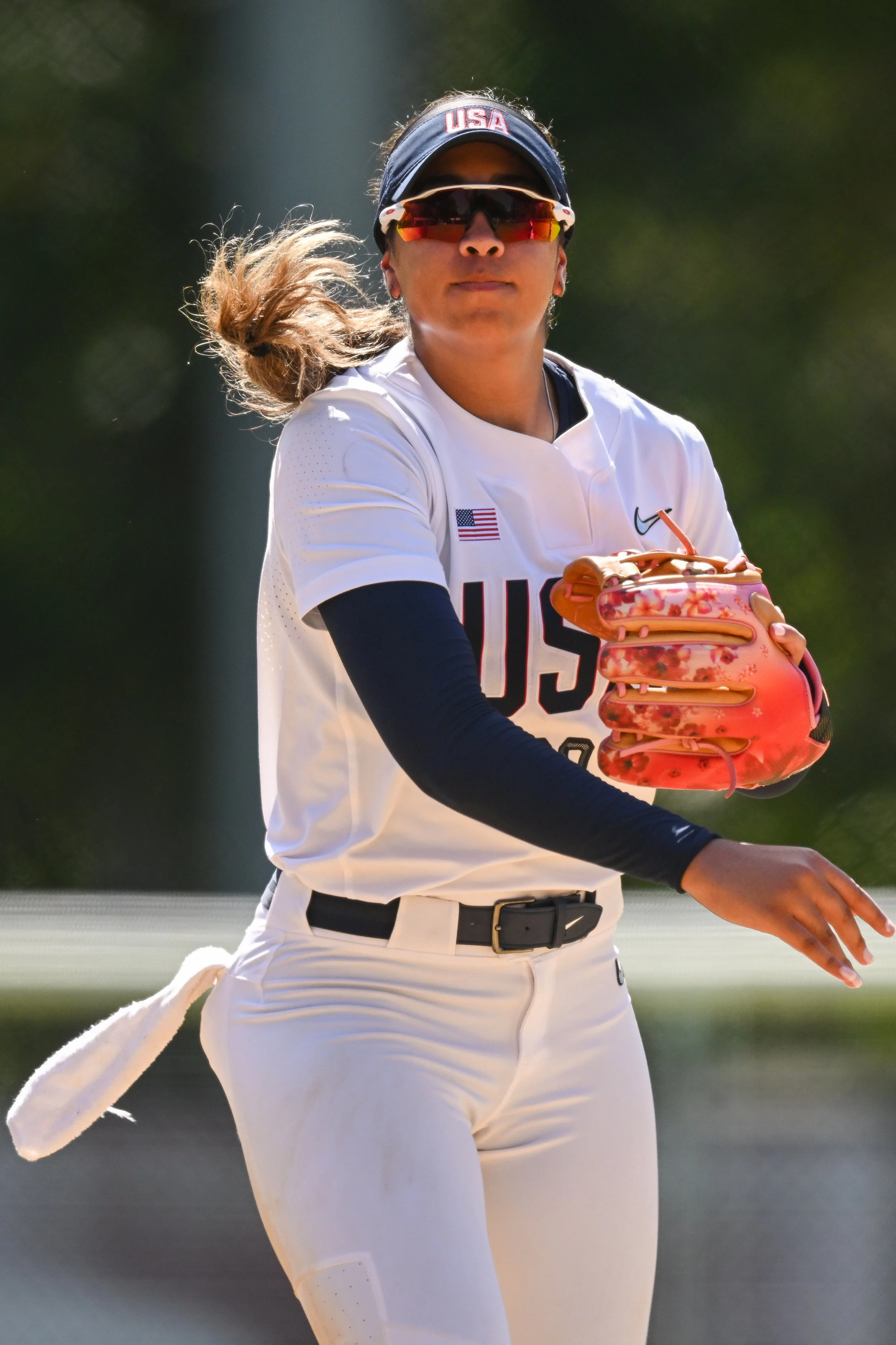 A female U.S. softball player in a white uniform with USA on the chest, wearing sunglasses and a headband, holding a pink softball glove, preparing for a play on the field.